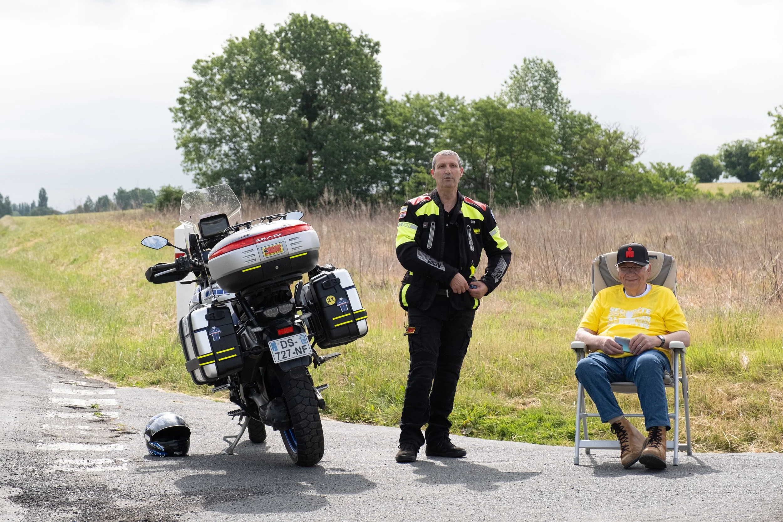 Un homme en uniforme de moto, un bénévole assis dans un fauteuil portant un t-shirt jaune, et une moto stationnée sur le bord d'une route de campagne avec des arbres en arrière-plan.