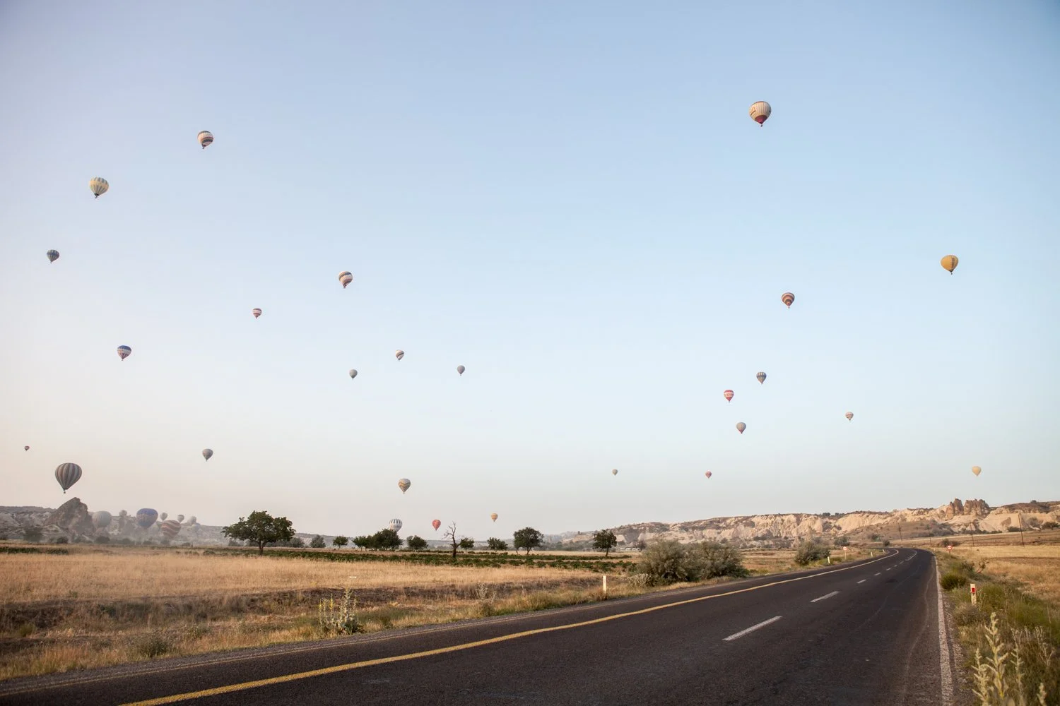 Ciel clair avec de nombreux ballons de baudruche colorés flottant au-dessus d'une route droite dans un paysage rural avec des collines et quelques arbres.