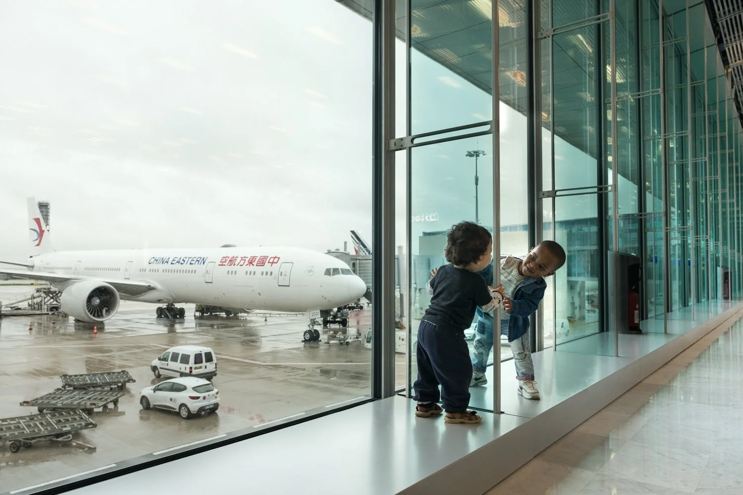 Deux enfants qui jouent à l'intérieur d'un terminal d'aéroport près d'une grande baie vitrée, avec un avion en dehors. Sur l'avion, il est écrit "China Eastern" en anglais et en chinois.