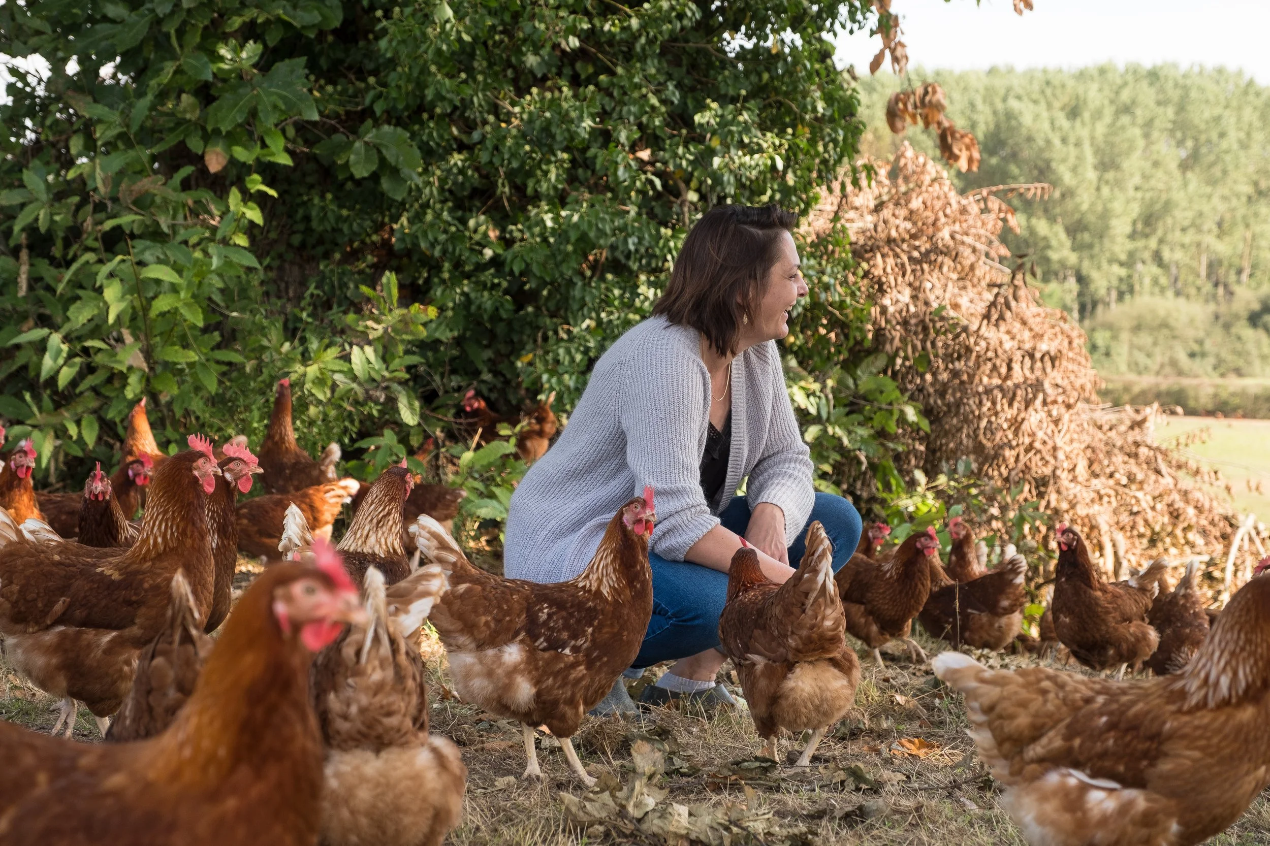Une femme souriante accroupie parmi des poules rousses dans un environnement extérieur avec des arbres et des buissons.