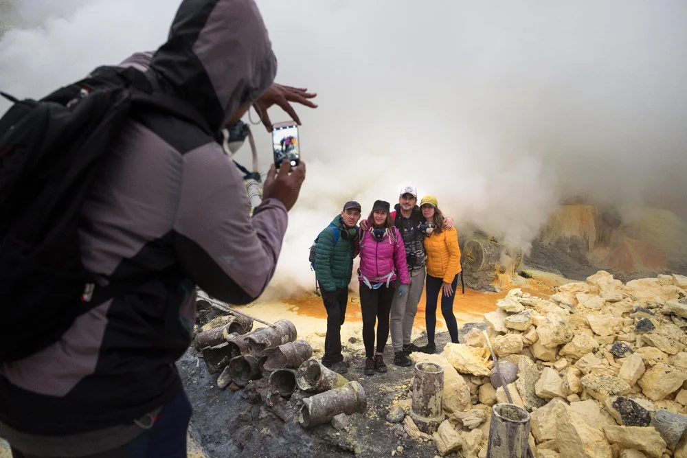 Groupe de quatre personnes en randonnée posant pour une photo devant une zone géothermique avec vapeur, roches jaunes et flammes dans un paysage volcanique.