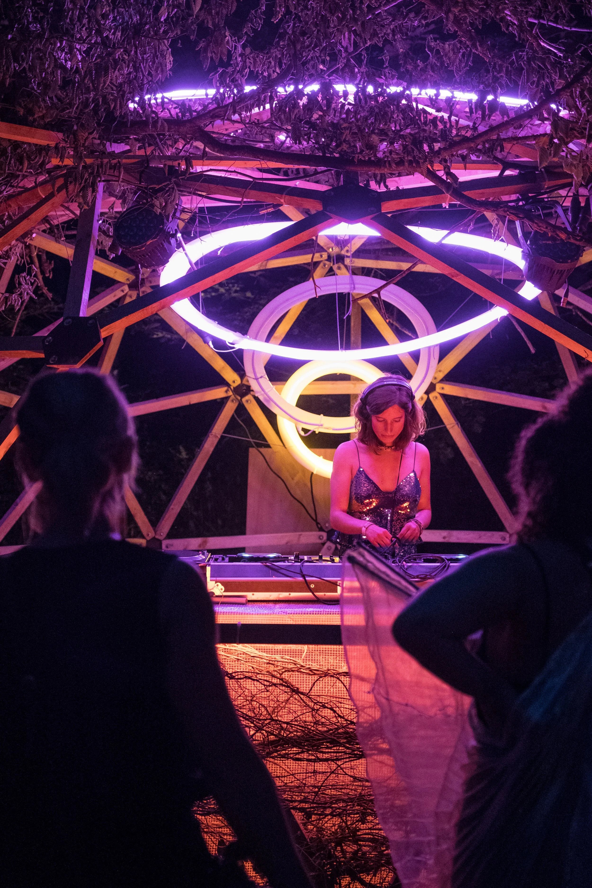 Une DJ joue de la musique lors d'un spectacle nocturne sous un décor de cercles lumineux violets et jaunes, avec un public regardant.