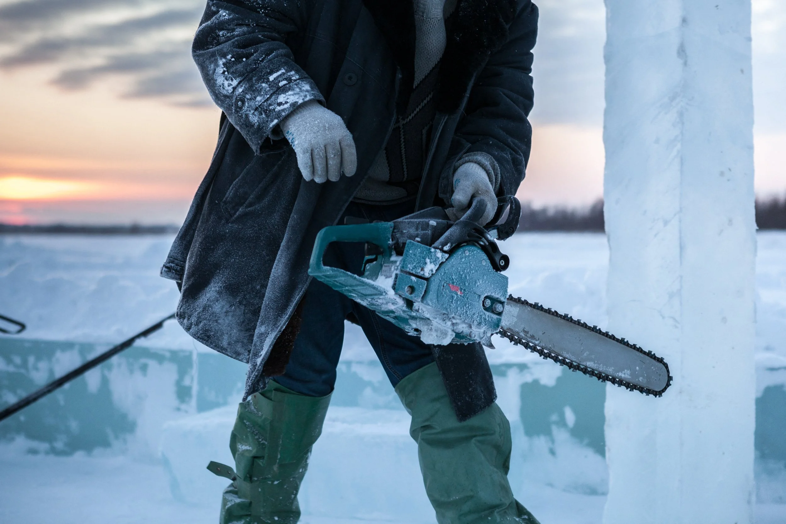 Personne utilisant une tronçonneuse électrique pour tailler de la glace dans un environnement froid en Russie, avec un coucher de soleil en arrière-plan.