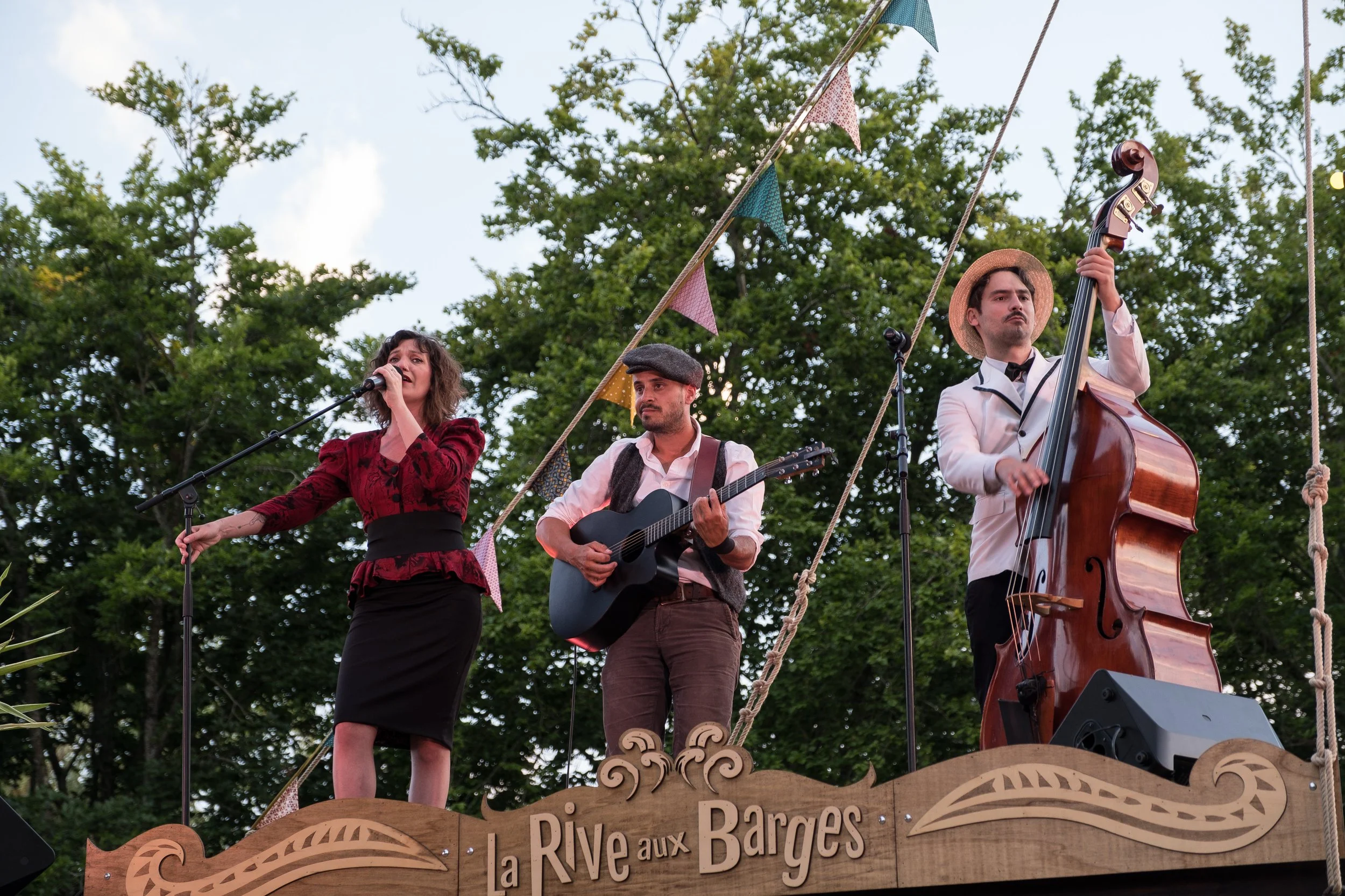 Un groupe de musiciens jouant sur une scène en bois décorée avec le panneau 'La Rive aux Barges', sous un ciel clair avec des arbres en arrière-plan, lors d'un spectacle en plein air.