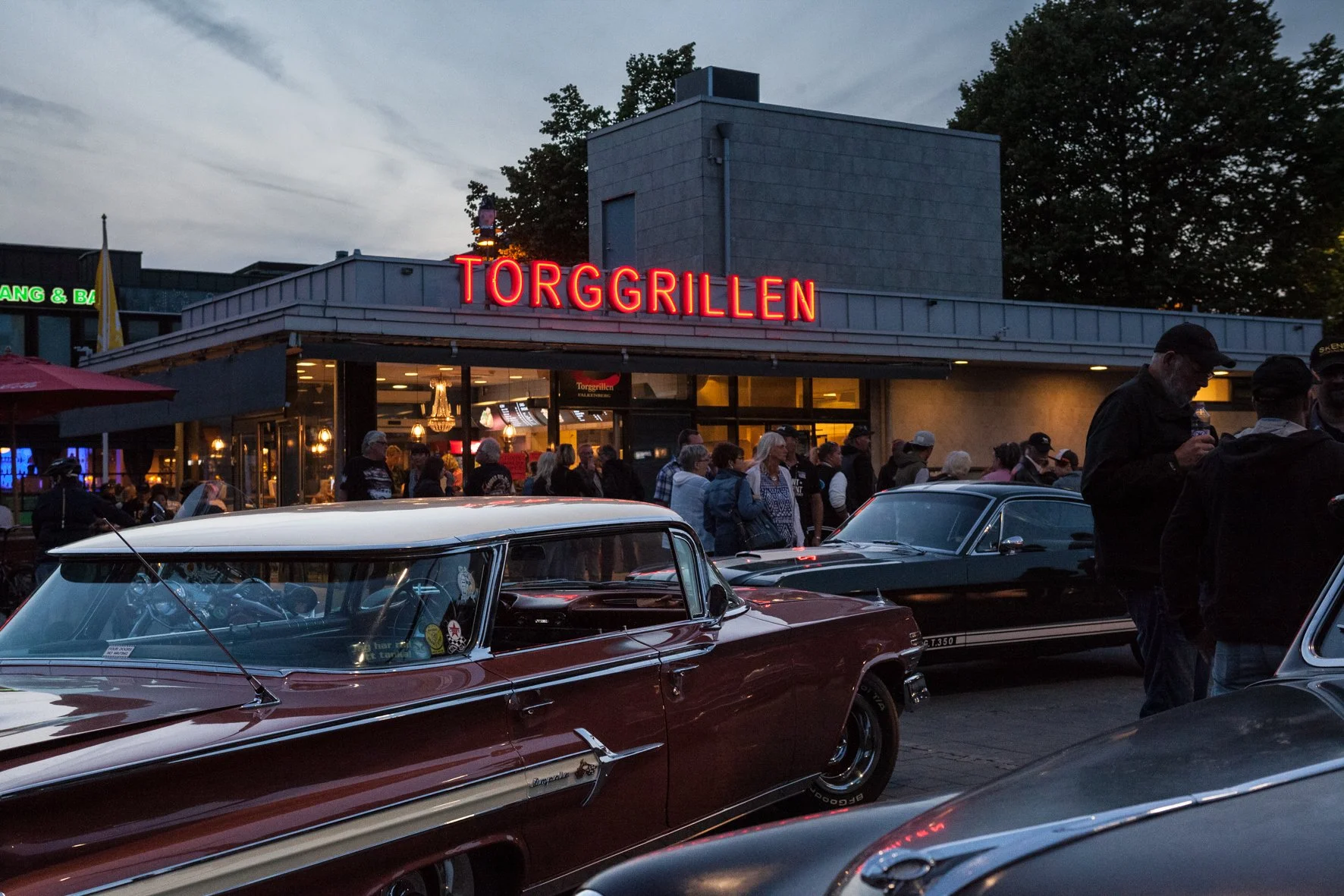 Une scène urbaine lors d'une soirée avec des voitures anciennes garées devant un restaurant appelé Torggrillen, où des personnes discutent et entrent dans l'établissement, sous un ciel crépusculaire en fin de journée.