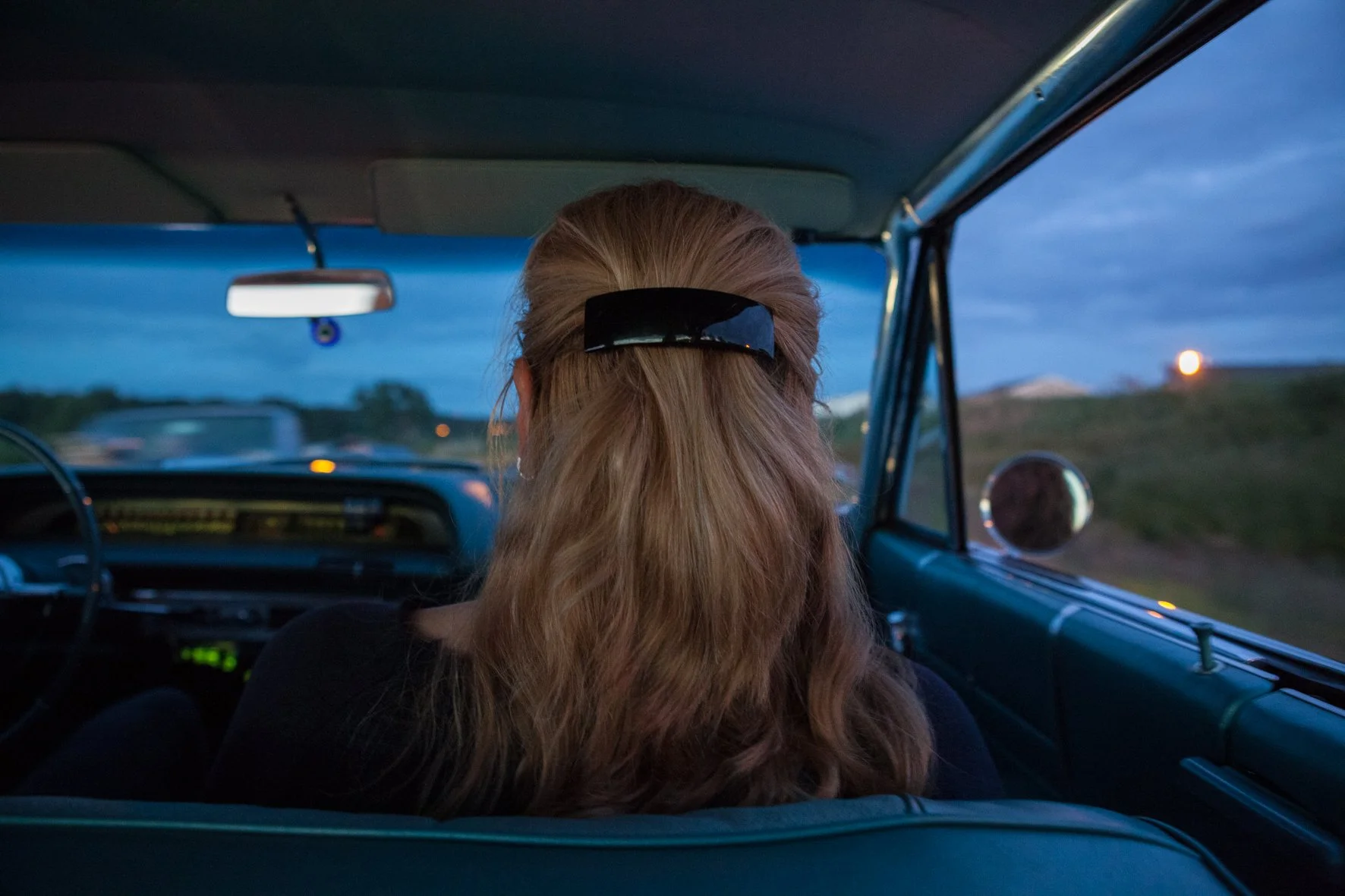 Vue à l'intérieur d'une voiture classique vue de l'arrière, avec une femme aux cheveux longs attachés par une pince noire, regardant par le pare-brise vers un ciel crépusculaire.