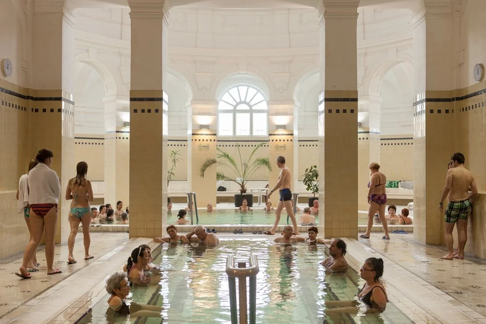 Piscine intérieure dans les bains Szechenyi à Budapest avec plusieurs personnes portant des maillots de bain, certaines flottant dans l'eau, d'autres discutant ou se relaxant sur le bord.