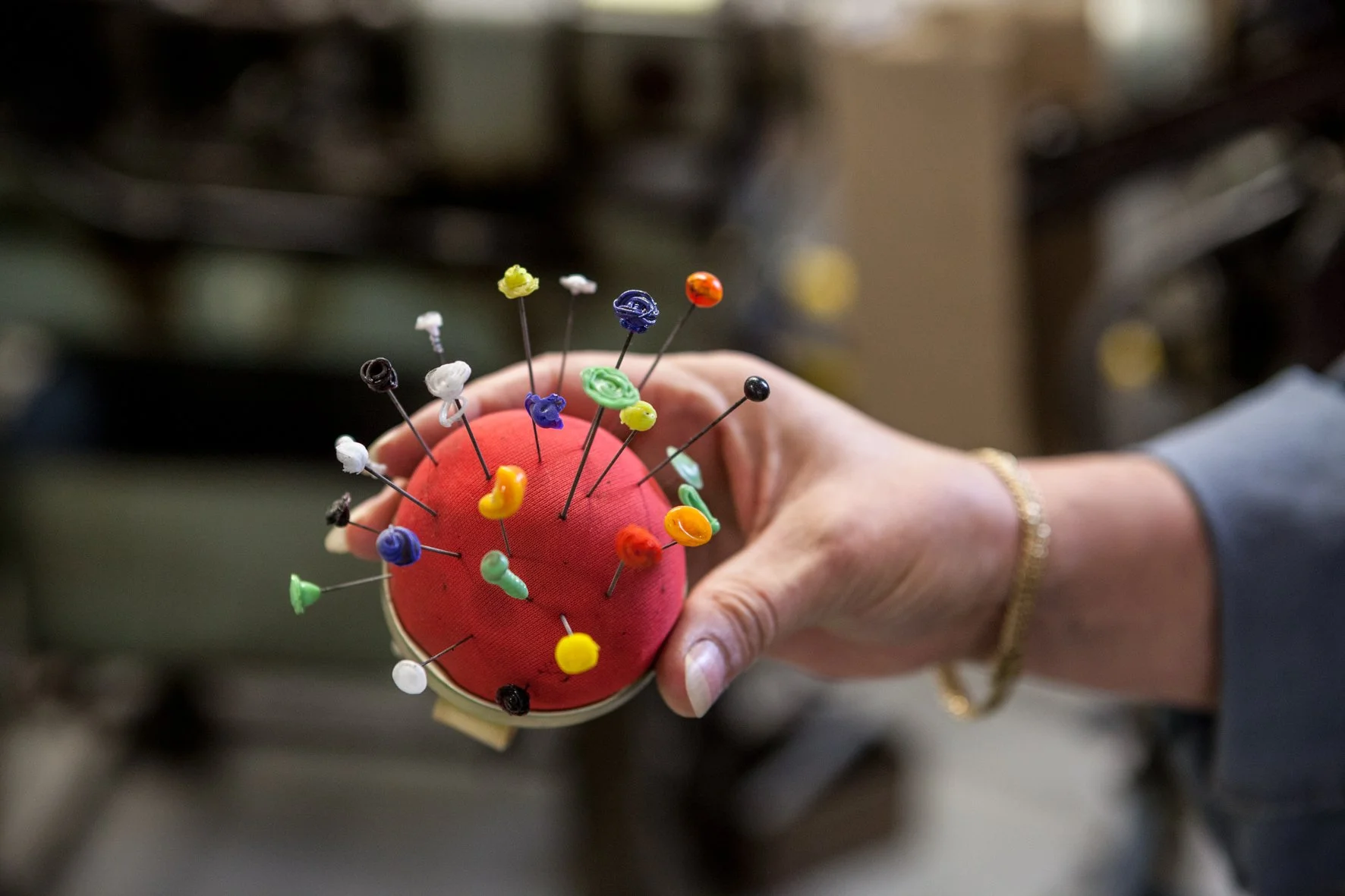 Une main tenant une boule de couture rouge avec des épingles multicolores piquées dans la boule.