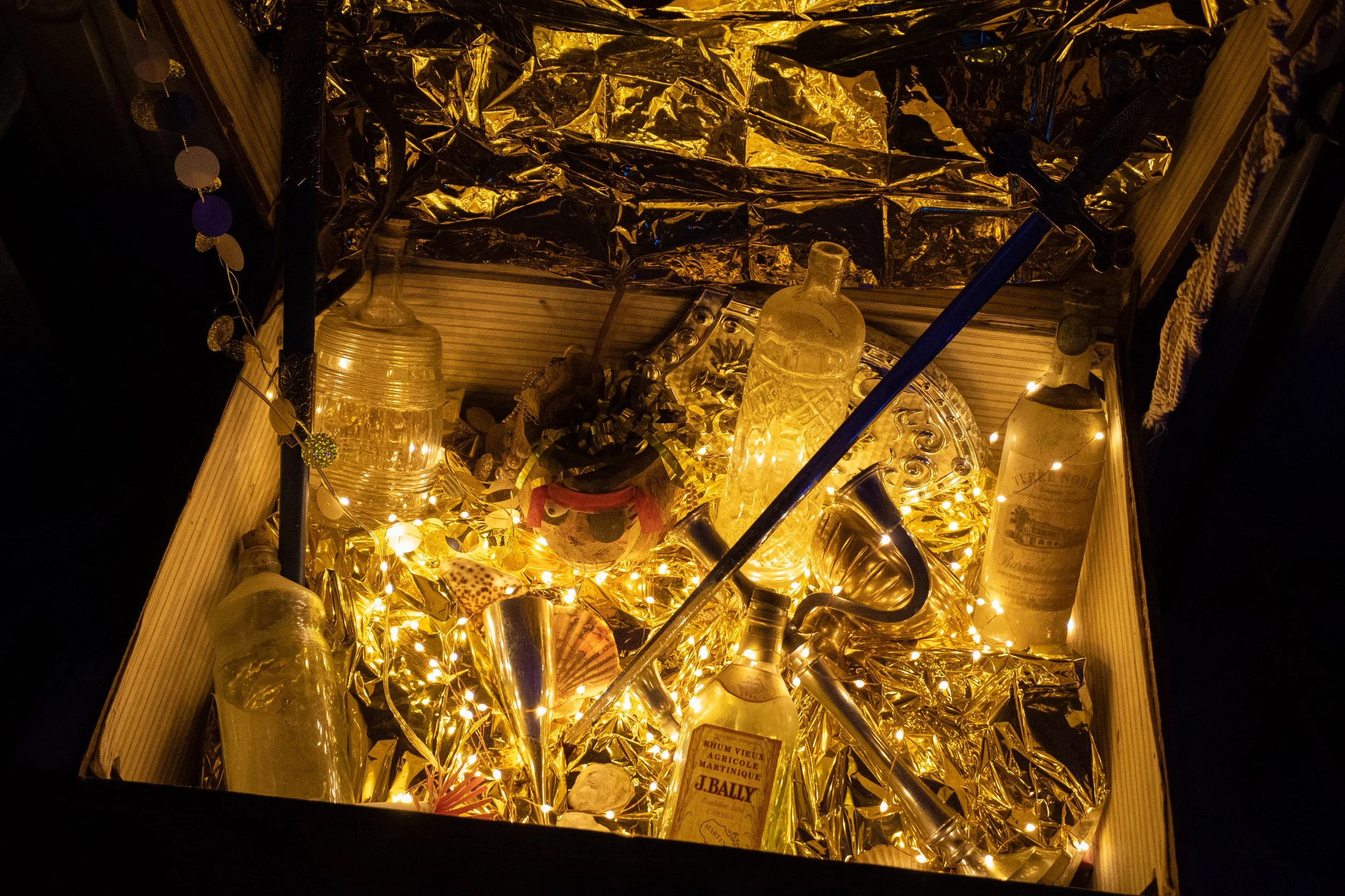 Une boîte en bois remplie de bouteilles en verre, d'une siphon, de décorations lumineuses dorées, et de papier d'aluminium.