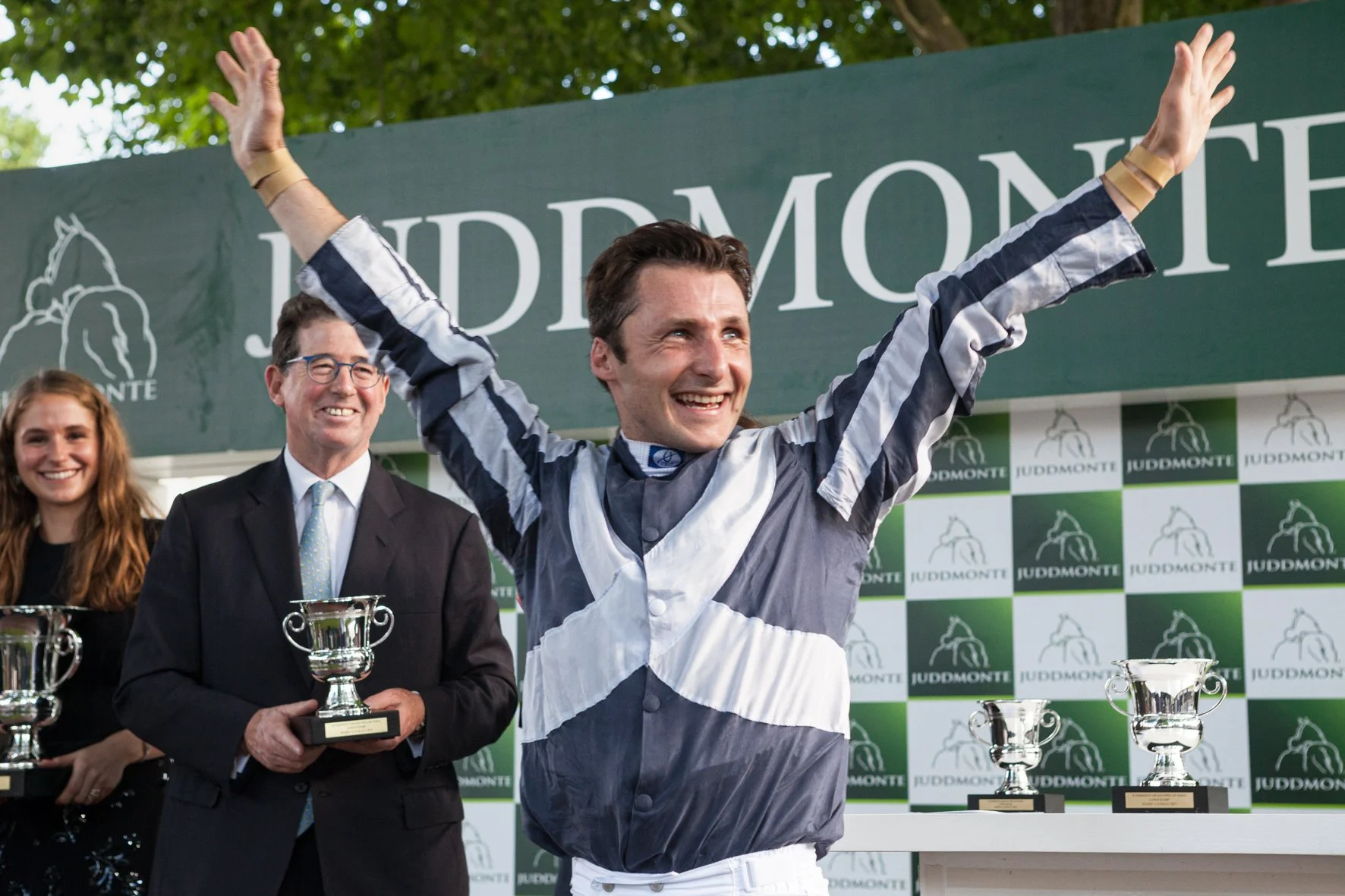 Un homme en tenue de jockey célèbre sa victoire avec ses bras levés en signe de triomphe, sur un podium de compétition.