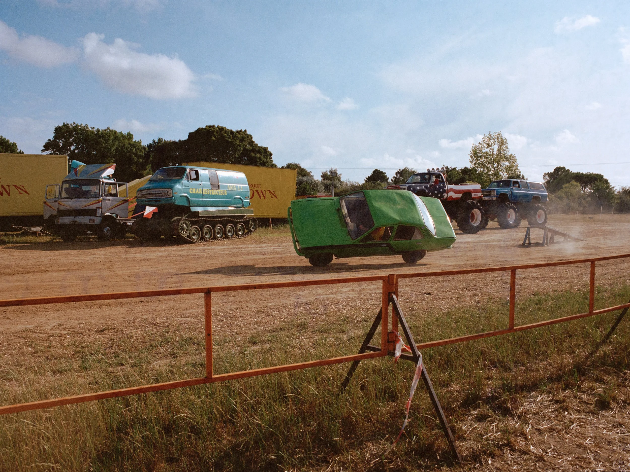 Une scène de course de véhicules déjantés, comprenant une voiture verte cabossée, un monstre truck coloré, un véhicule avec des ailes, et d'autres voitures, sur un terrain poussiéreux avec un ciel partiellement nuageux.