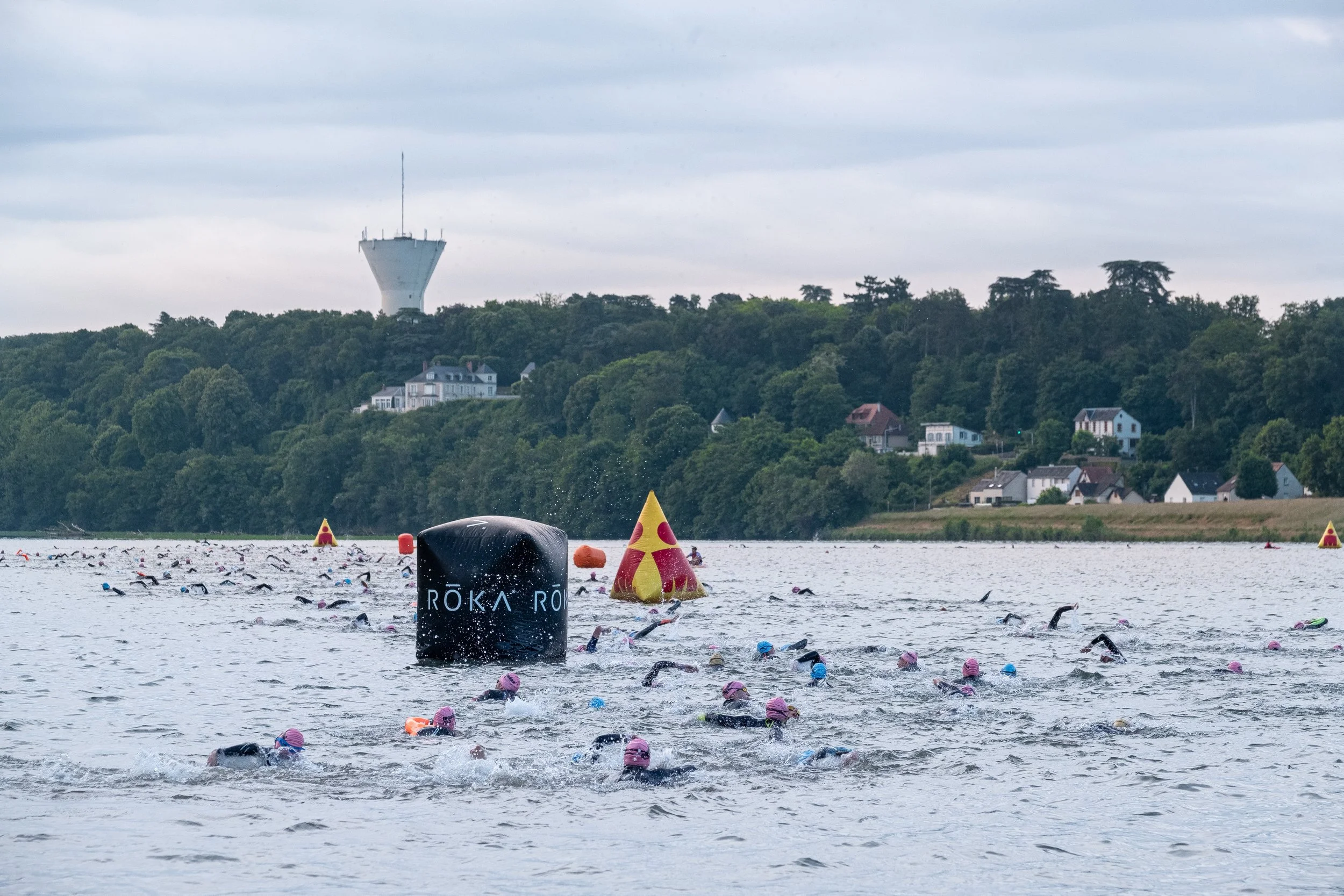 Nombreux triathlètes nageant dans une rivière avec des bouées et des totems de course, un paysage verdoyant en arrière-plan avec des maisons et une tour de communication.