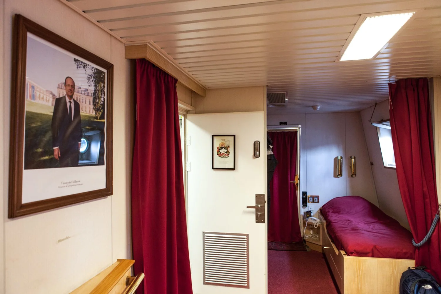 Cabine de bateau avec lit simple sur la frégate militaire Surcouf. Fenêtres, rideaux rouges, tableau encadré sur le mur, porte avec grille d'aération, et accessoires tels que téléphones et lampes.