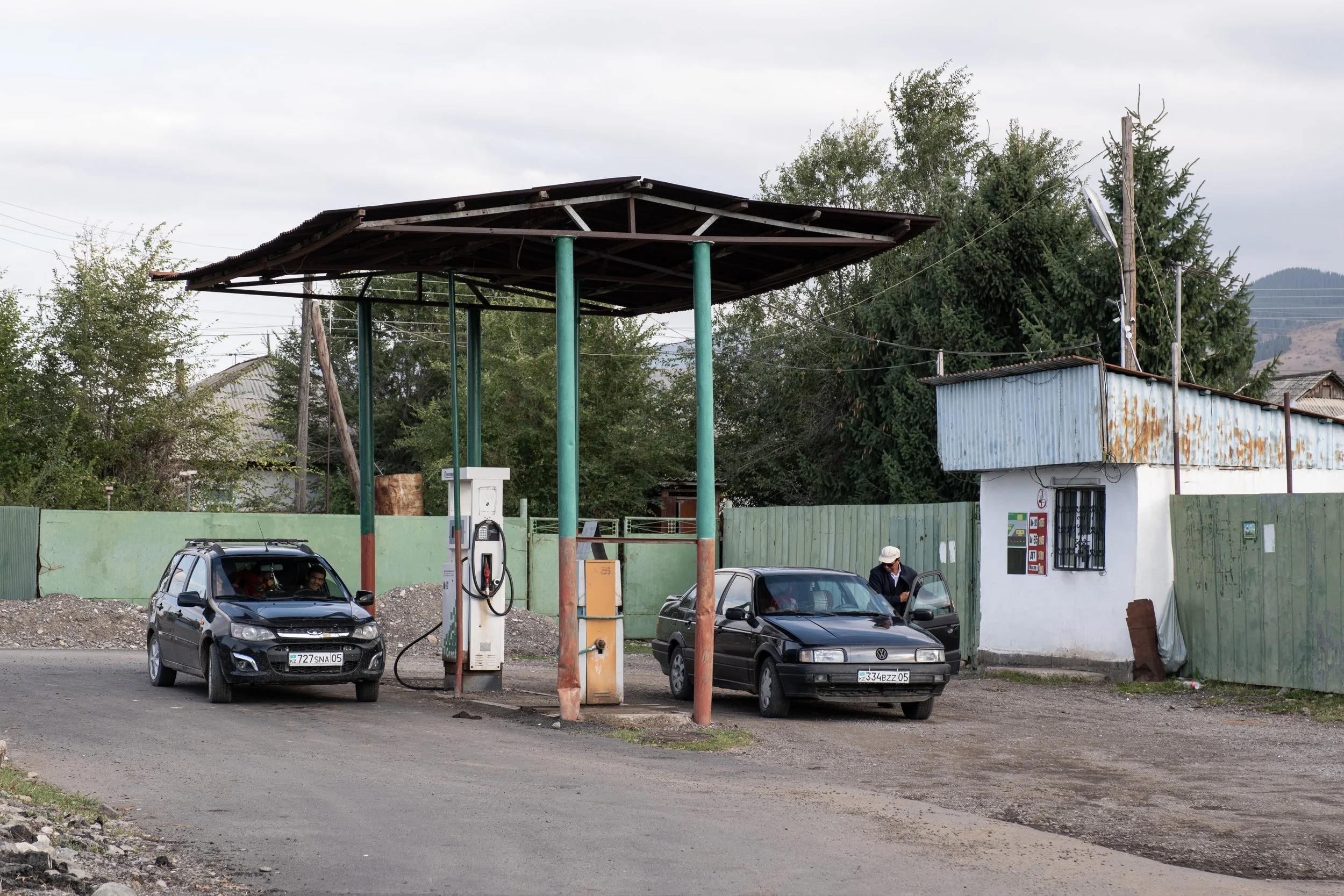 Station-service avec deux voitures, une personne faisant le plein, bâtiment en arrière-plan, paysage rural, au Kazakhstan.