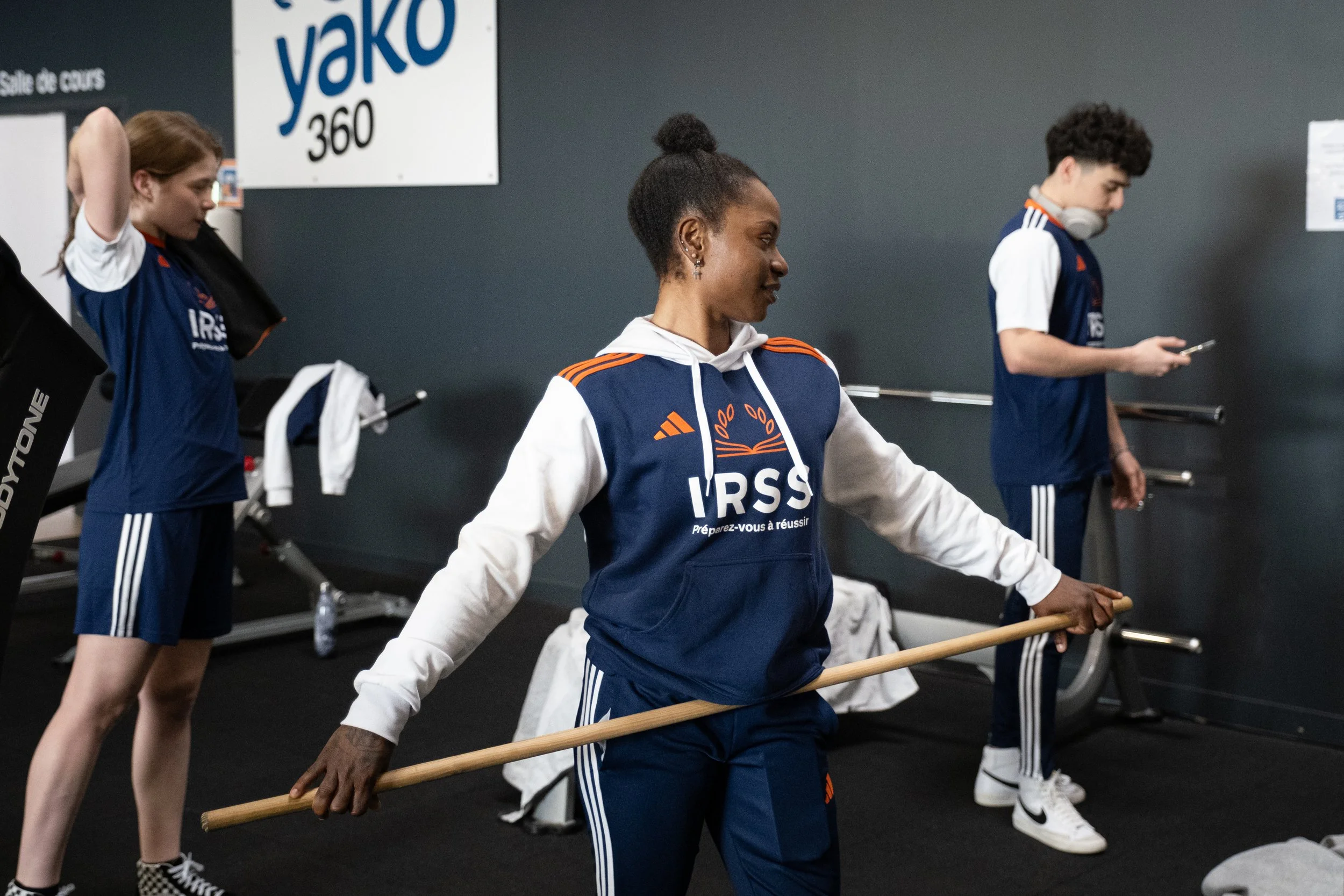 Trois jeunes sportifs en tenue d'entraînement dans une salle de sport, avec une femme au centre utilisant une arme de gymnastique pour l'entraînement, et des deux autres utilisant leurs téléphones. Logo 'yako 360' visible en arrière-plan.