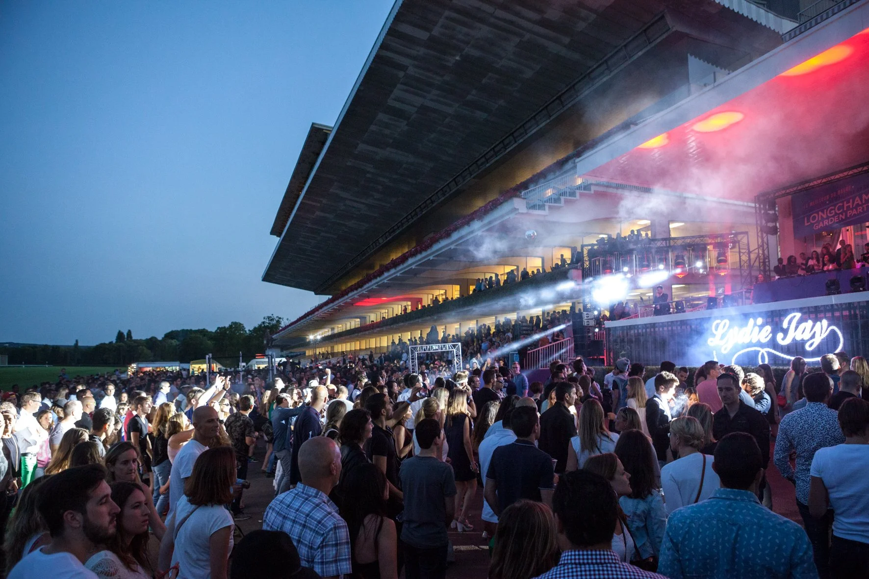 Une foule de personnes assiste à un concert en plein air à l'hippodrome de Longchamp, avec un éclairage coloré, en soirée.