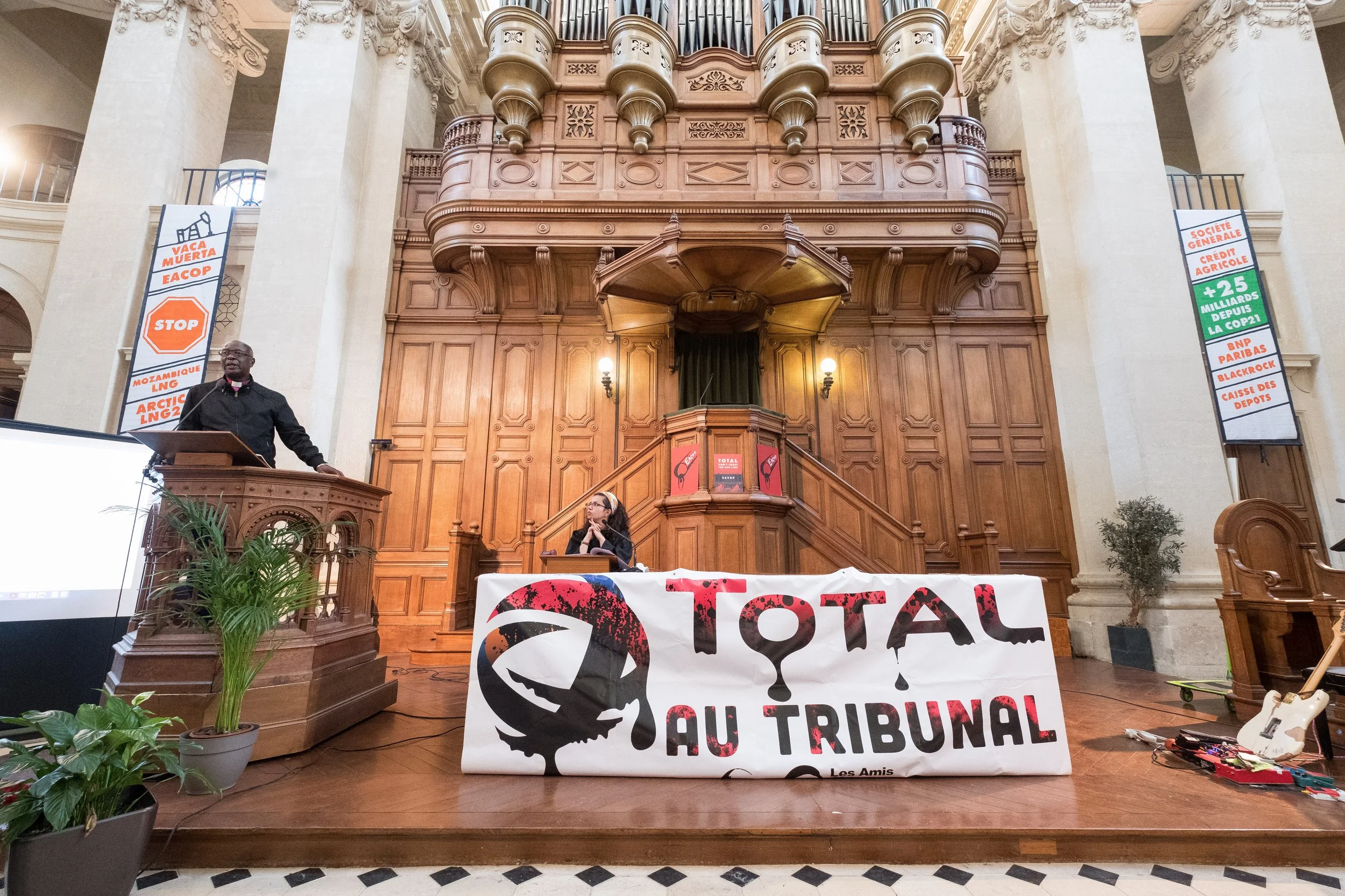 Une salle de tribunal avec un panneau de protestation sur la table, des bannières avec des messages sur le changement climatique et la justice. Deux personnes sont présentes, l'une parle à un micro et l'autre est assise à une table.