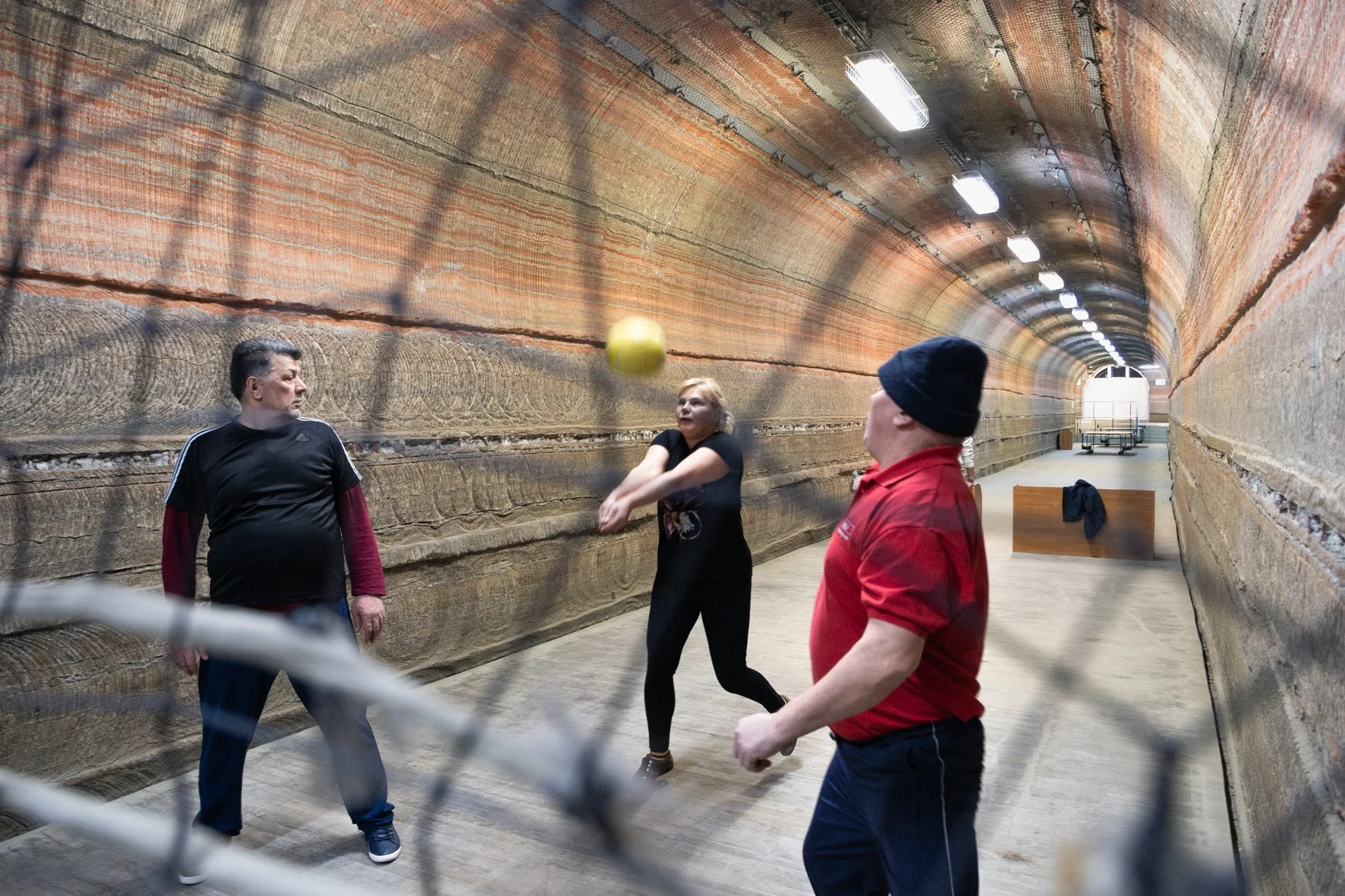 Trois personnes jouent au volley-ball dans un tunnel avec murs en pierre rouge, éclairé par des lampes suspendues.