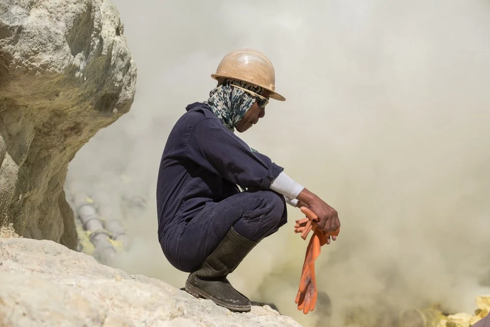 Mineur en combinaison de travail, portant un casque en plastique, des lunettes de protection, et tenant une paire de gants en caoutchouc orange, à proximité d'une zone de fumée ou de vapeur.