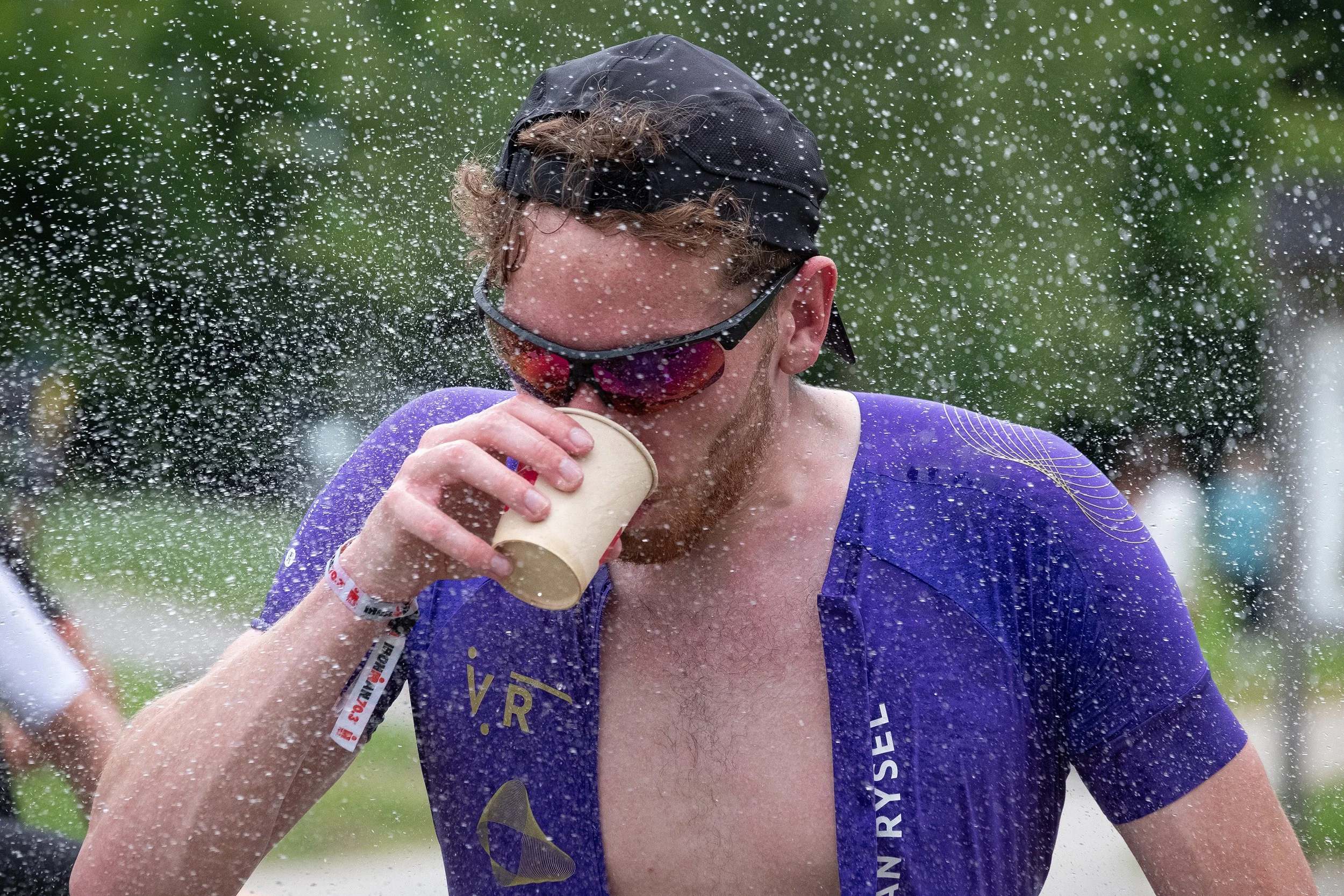 Un homme portant un maillot violet, une casquette noire et des lunettes de soleil est en train de boire dans un gobelet en papier lors d'une compétition ou d'un événement en plein air. Il est entouré de gouttelettes d'eau.
