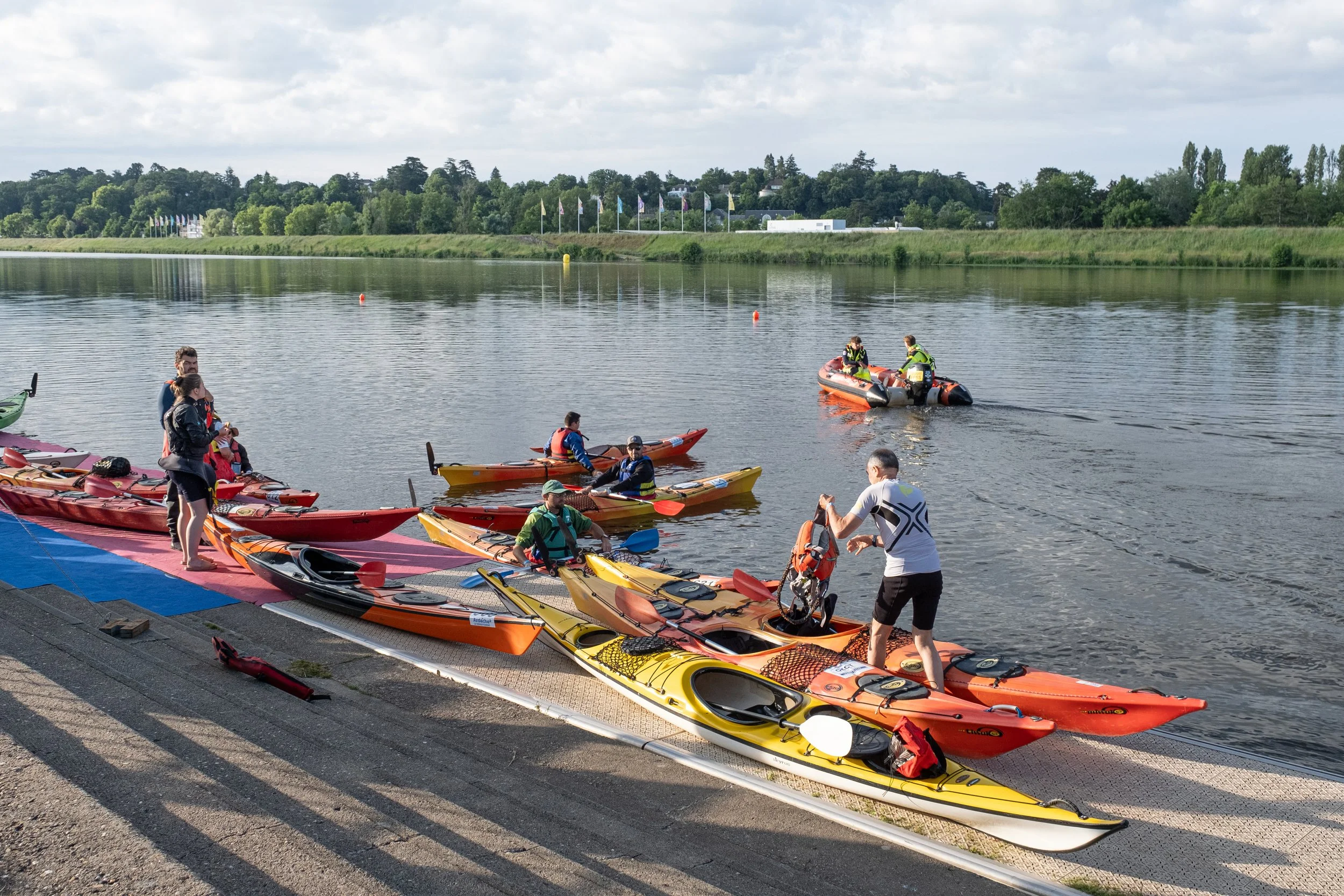 Groupe de personnes préparant des kayaks colorés au bord de l'eau, avec quelques personnes en train de pagayer et des policiers en bateau de secours au loin.