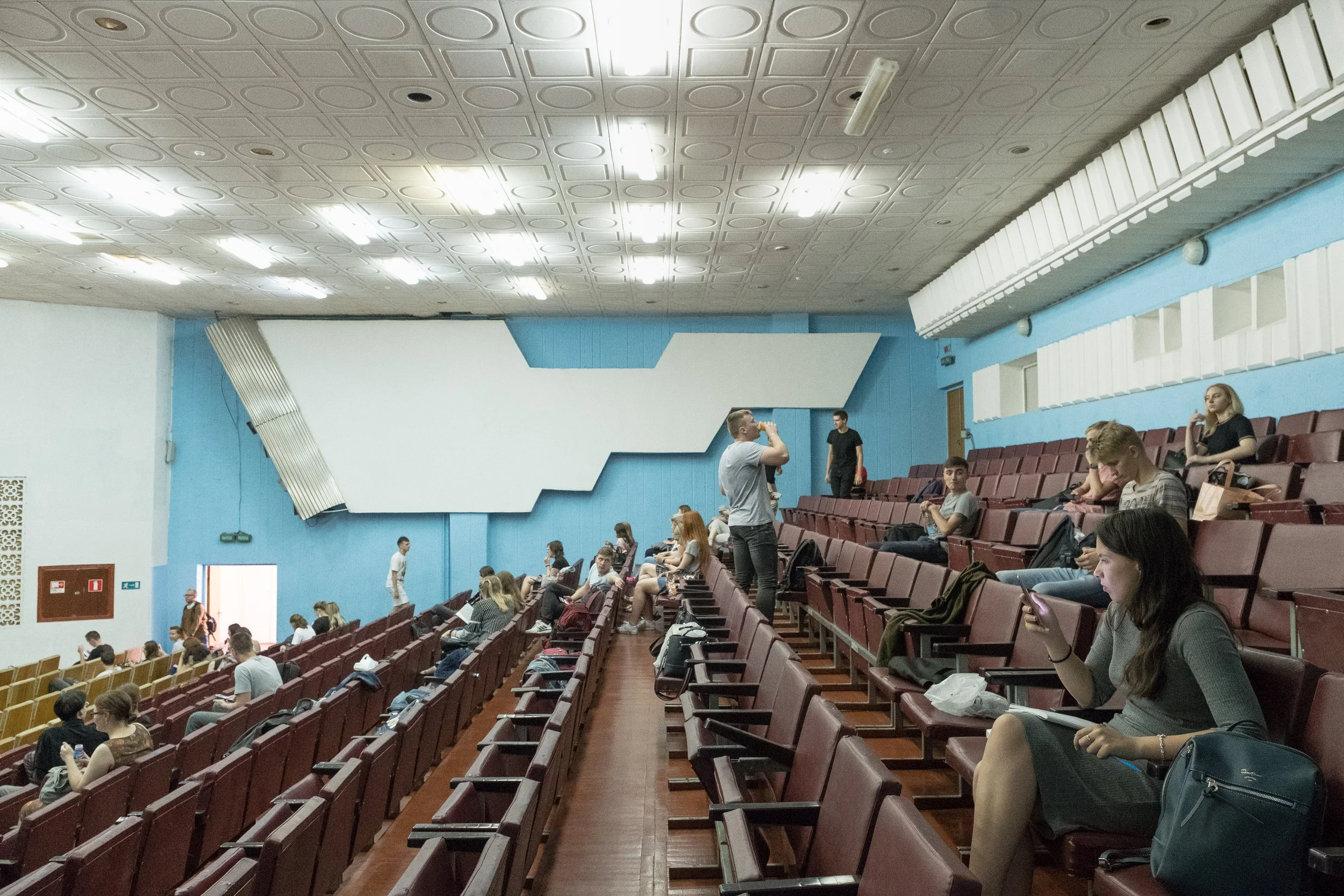 Amphithéatre universitaire avec des sièges rouges à Gomel, en Biélorussie, quelques personnes attendent que le cours commence. Le plafond est doté de lumières fluorescentes, murs bleus et une grande structure blanche sur le mur.