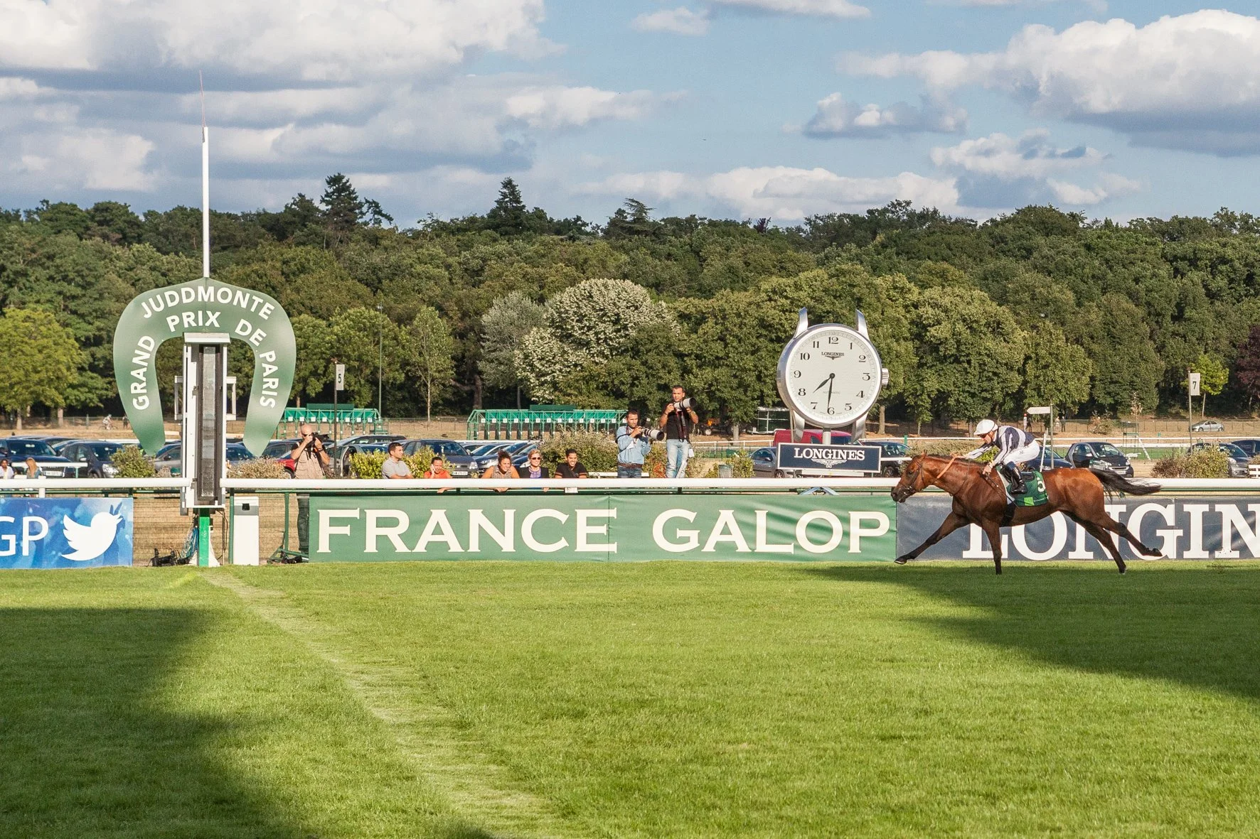 Course hippique à l'hippodrome de Longchamp avec un jockey à cheval sur la piste verte, panneaux publicitaires, un horloge géante et un groupe de personnes regardant la course, dans un environnement naturel avec des arbres et un ciel nuageux.