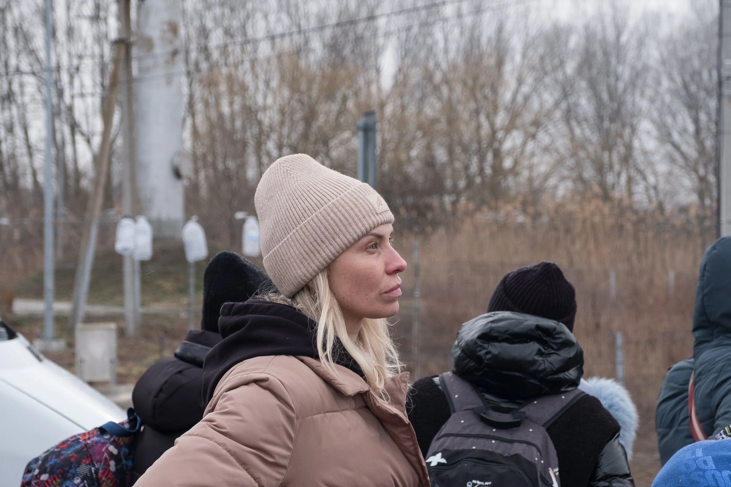 Une ukrainienne portant un bonnet beige et une veste marron, observant quelque chose au loin parmi un groupe de réfugiées vêtues de vêtements d'hiver dans un environnement extérieur avec des arbres sans feuilles et un ciel nuageux.