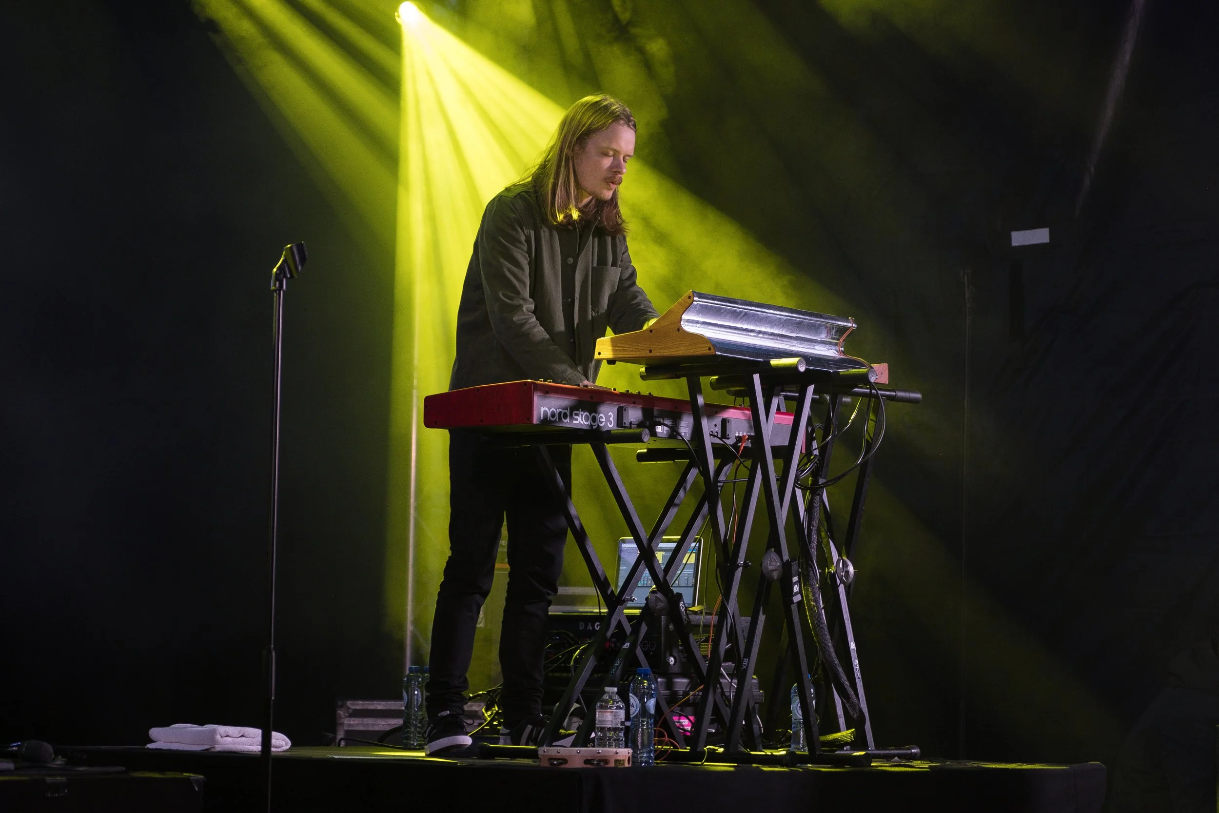 Un musicien avec des cheveux longs joue du clavier sur scène, éclairé par des lumières jaunes.