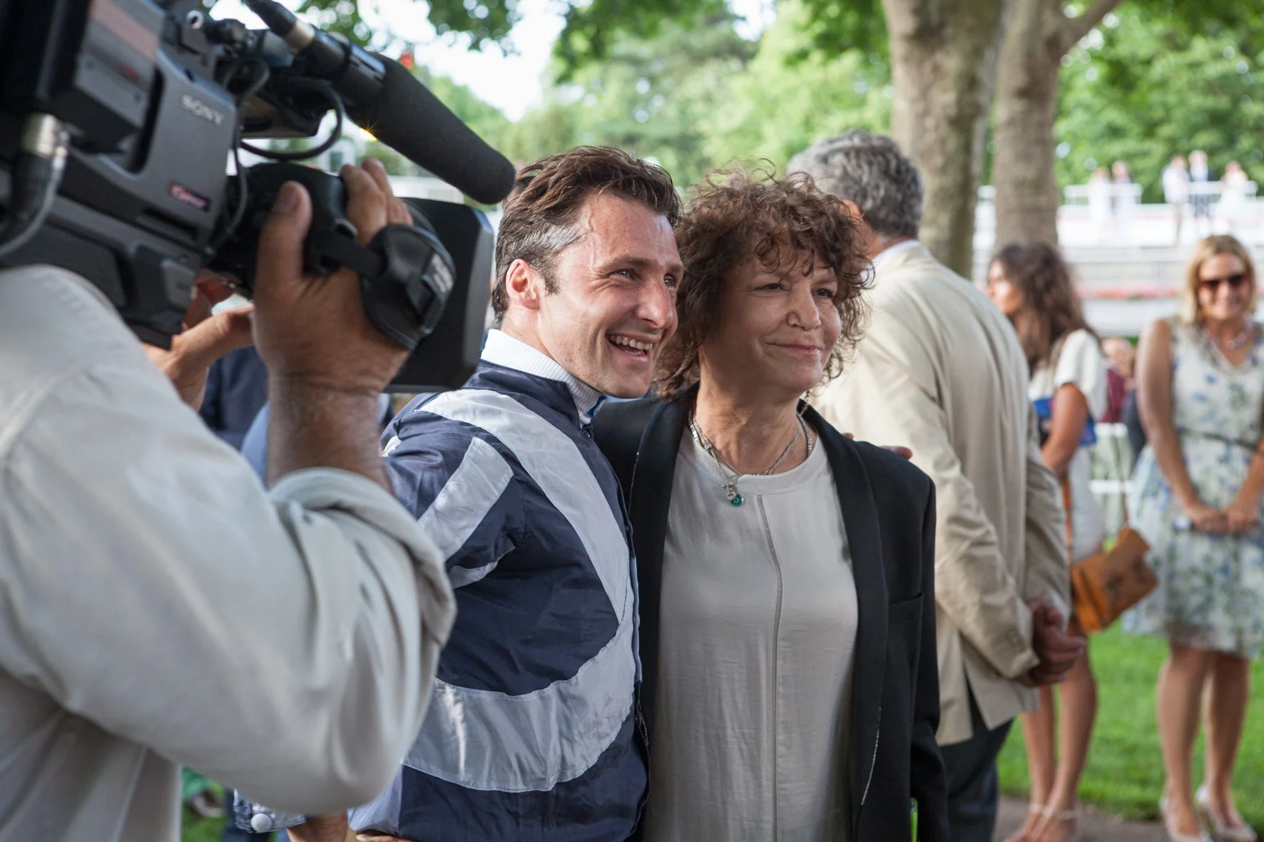 Deux personnes posent et sourient pour la caméra lors d'une course hippique, avec d'autres personnes en arrière-plan.