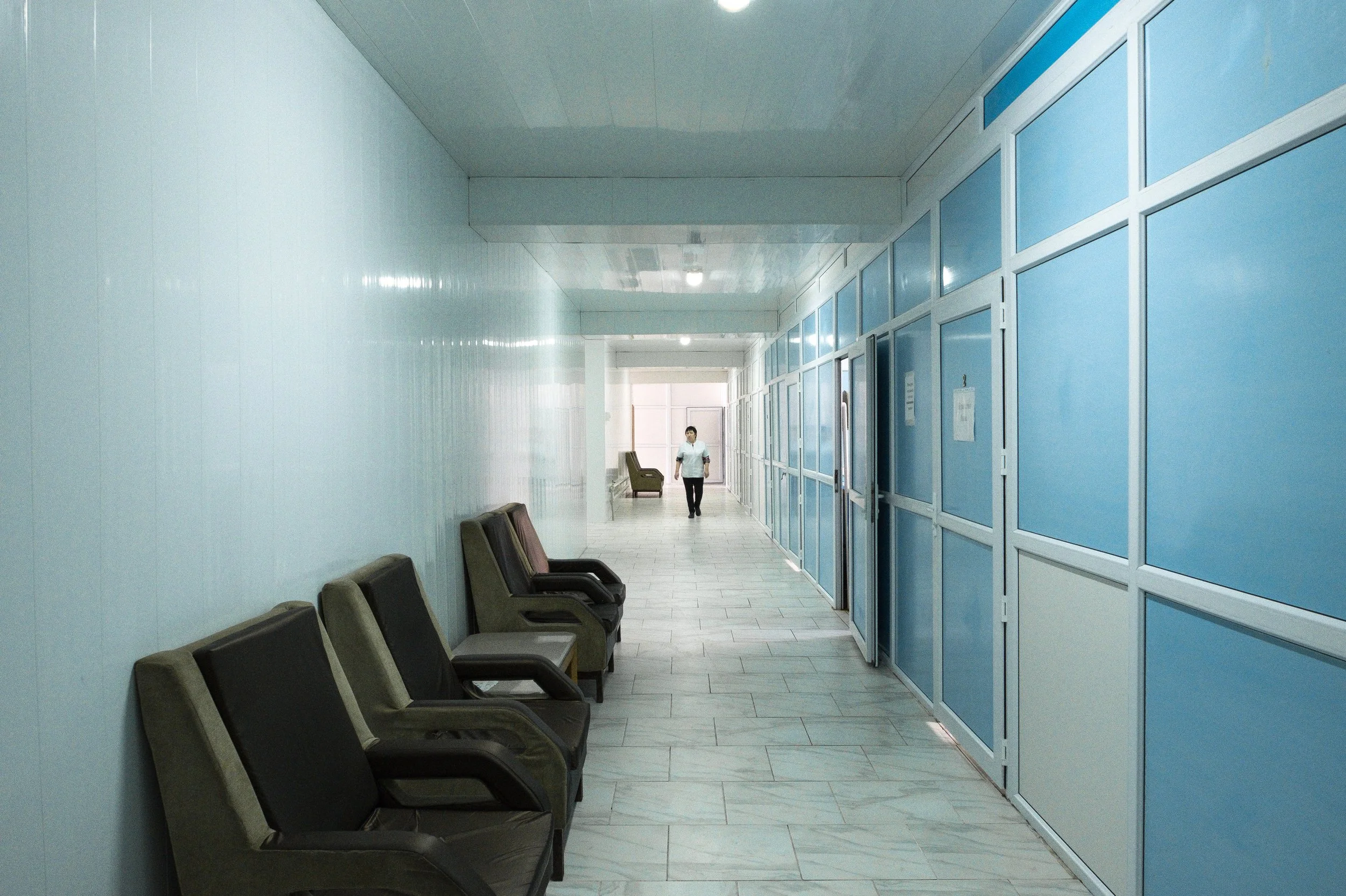 Couloir d'un sanatorium avec plusieurs fauteuils sur la gauche et un homme portant un masque marchant au centre, à l'arrière une porte de sortie.