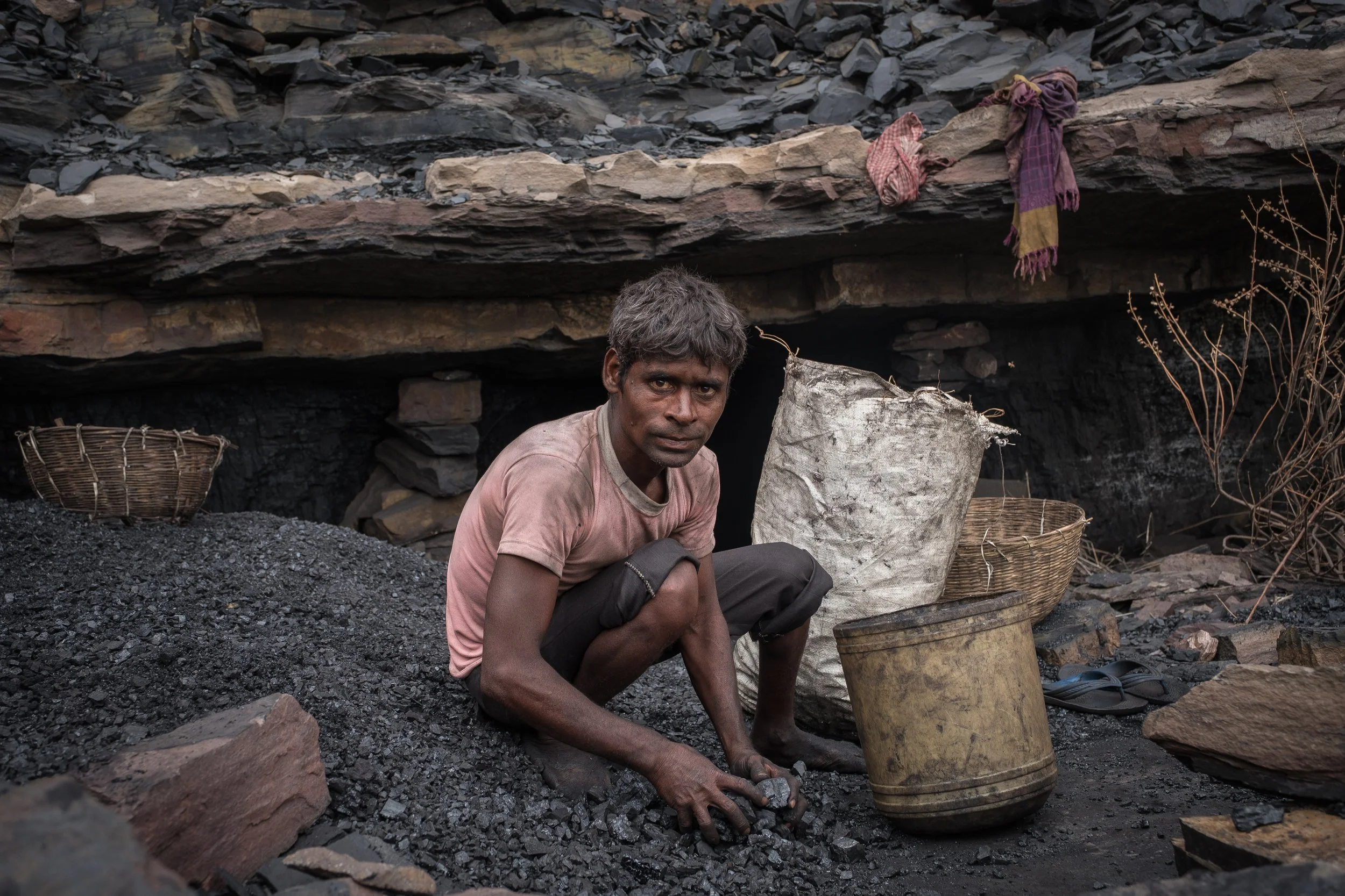 Un homme dans une mine, entouré d'outils et de sacs, avec des vêtements sales, sous un abri rocheux.