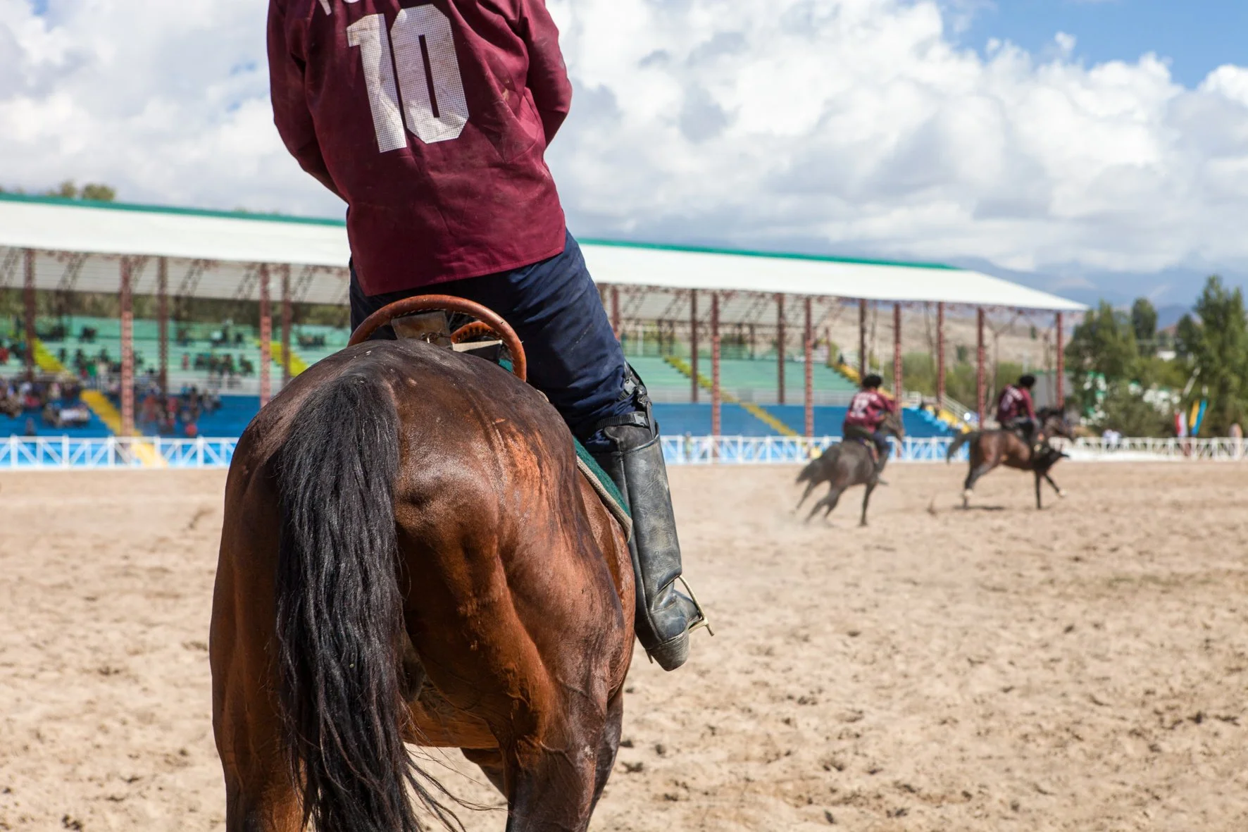 Personne à cheval portant un maillot rouge avec le numéro 10, vue partielle du dos, en première plan. En arrière-plan, plusieurs autres cavaliers participent à un match de kok boru sur un terrain en terre, sous un ciel partiellement nuageux.