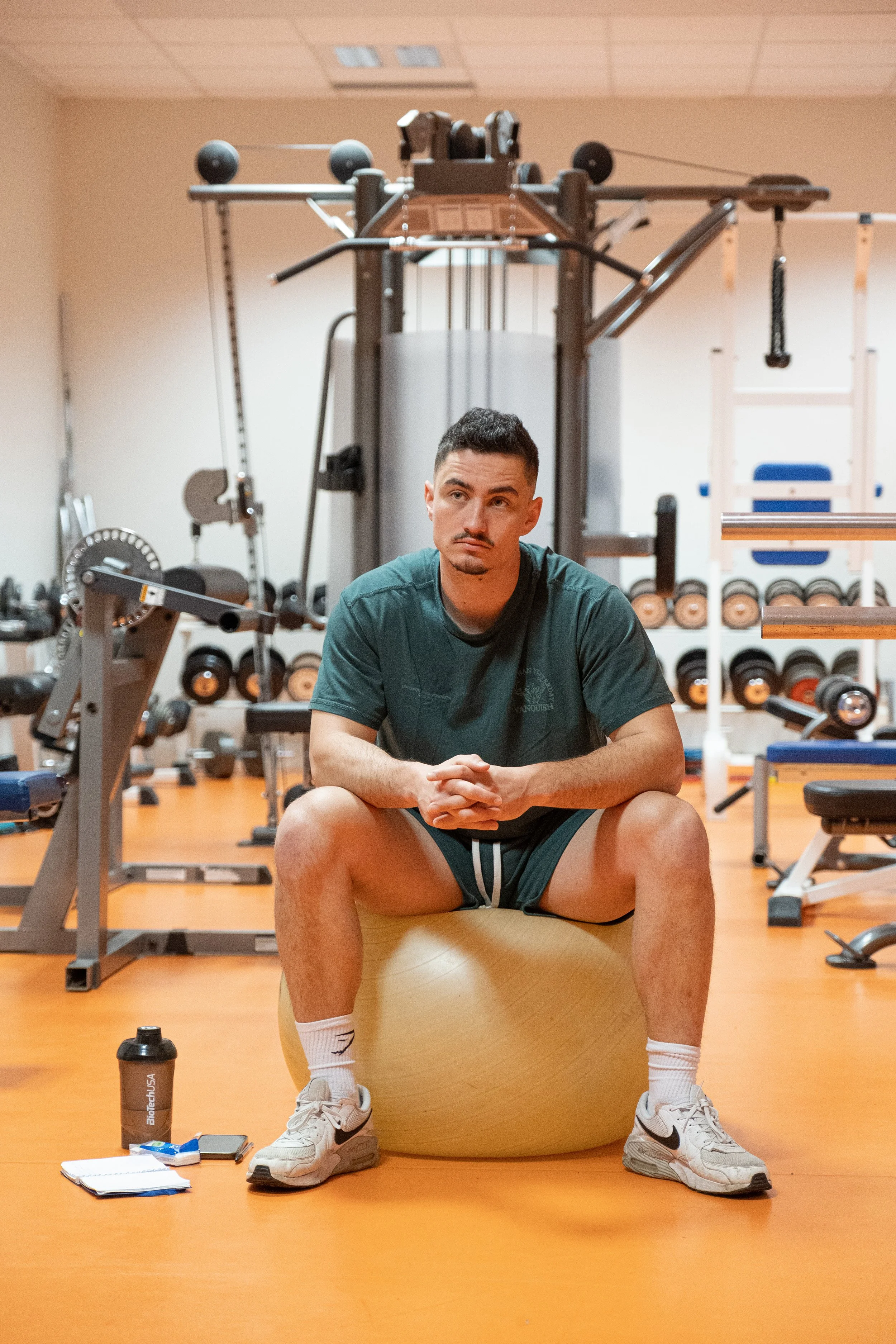 Un homme jeune assis sur un ballon d'exercice dans une salle de gym, entouré d'équipements de musculation et de poids.
