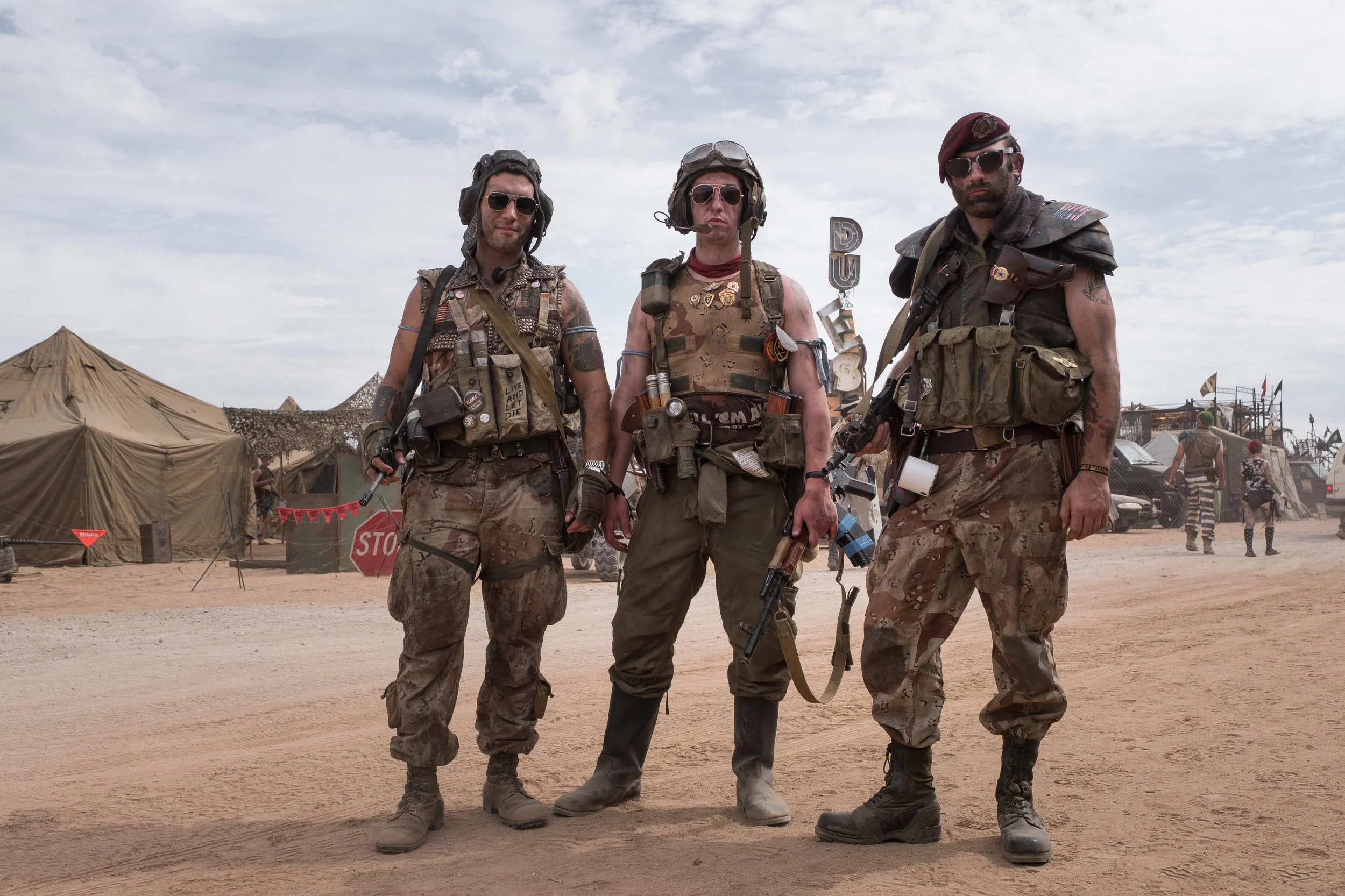 Trois hommes en tenue militaire sale, portant des lunettes de soleil, casques, et avec des équipements, se tiennent dans un camp désertique.