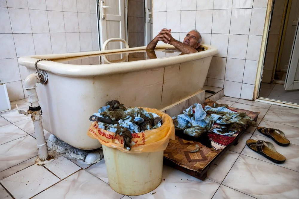 Un homme âgé dans une baignoire, dans une salle de bain avec des déchets et des vêtements sales autour.