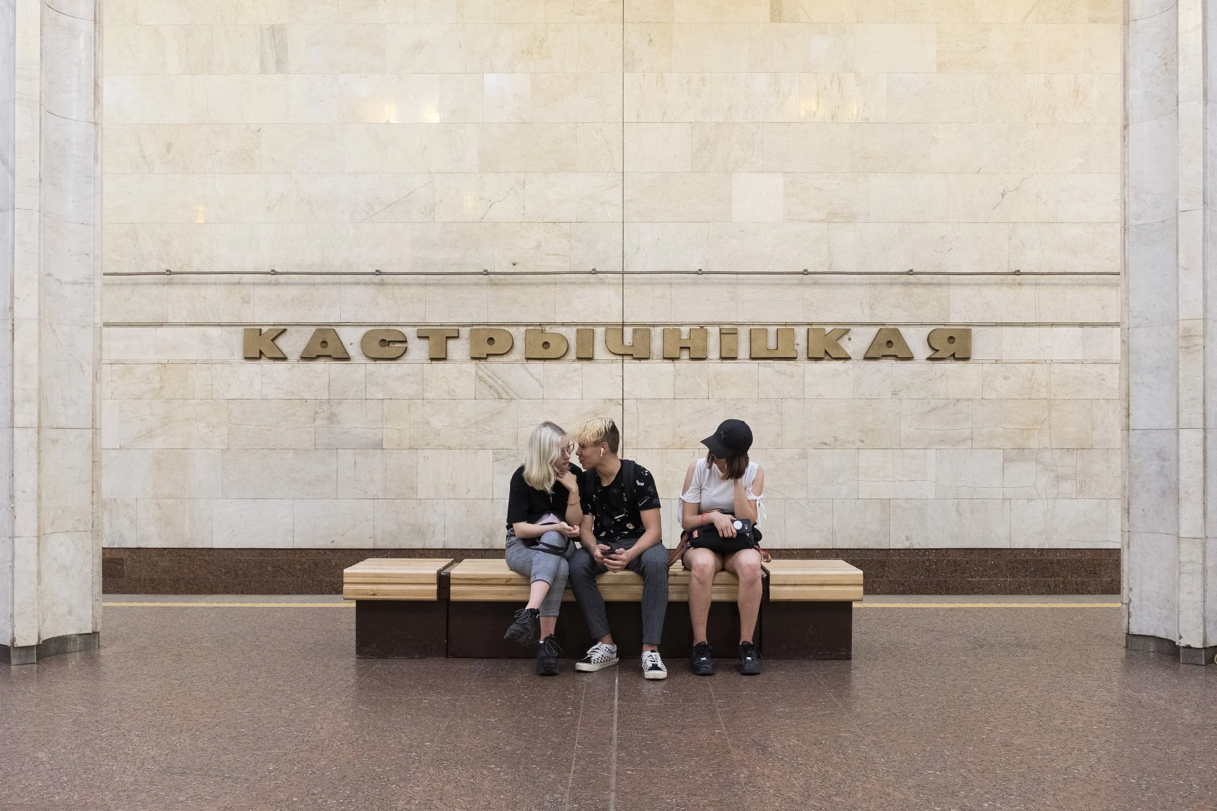 Trois jeunes assis sur un banc dans une station de métro en Biélorussie. Le mur derrière eux a une inscription en cyrillique qui dit 'Кастрычніцкая'.