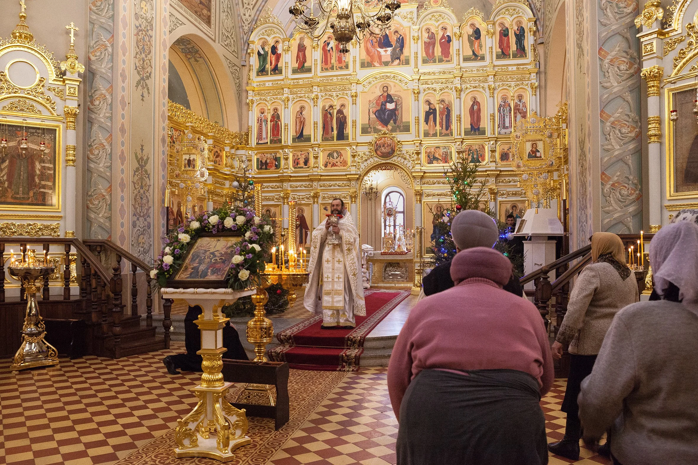 Une cérémonie religieuse dans une église orthodoxe, avec un prêtre en costume blanc devant l'autel orné d'icônes et de bougies, des personnes priant, un arbre de Noël décoré, et une décoration riche en dorures.