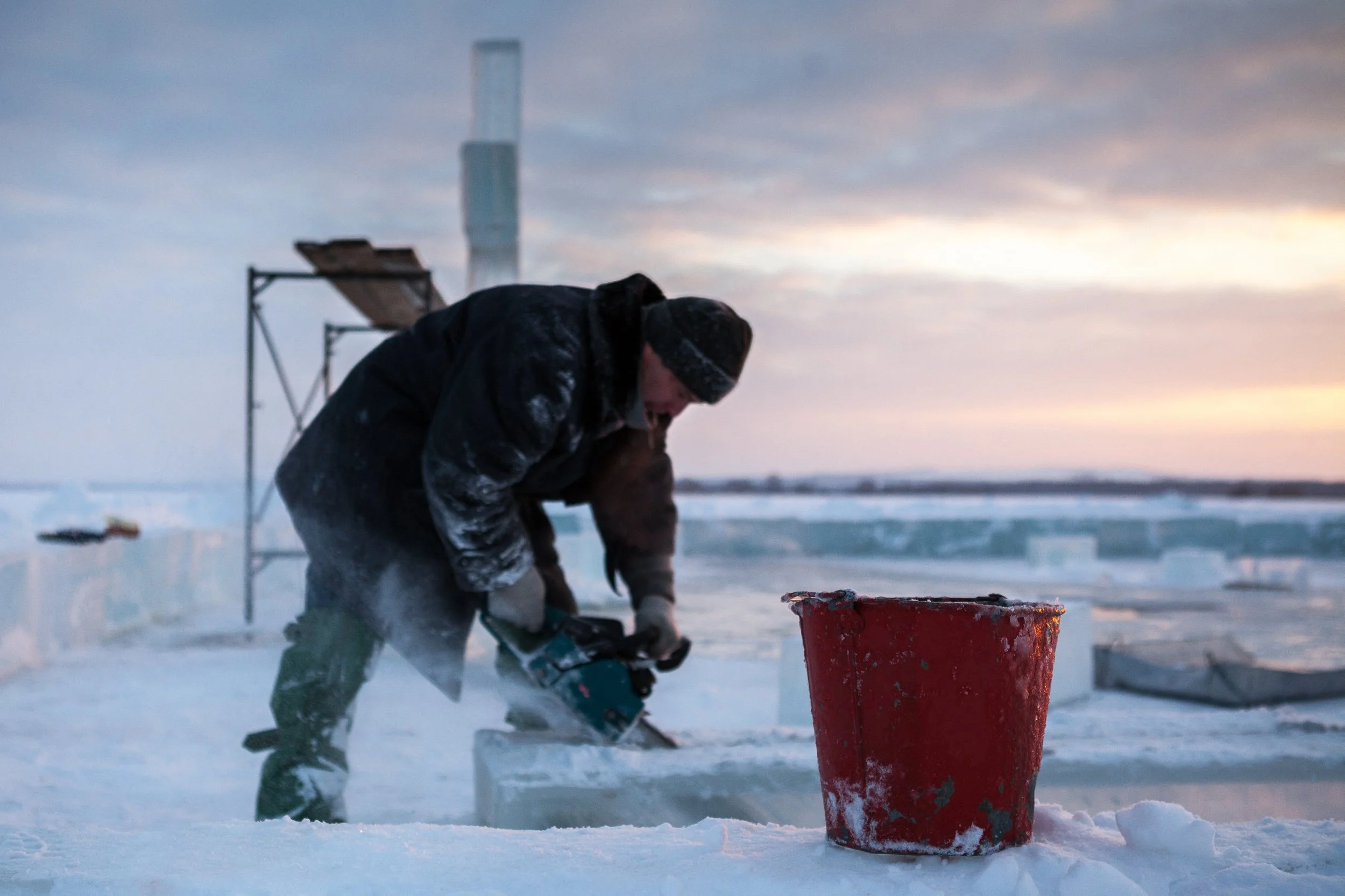 Une personne coupe de la glace dans un paysage enneigé lors du coucher du soleil avec un seau rouge à proximité.