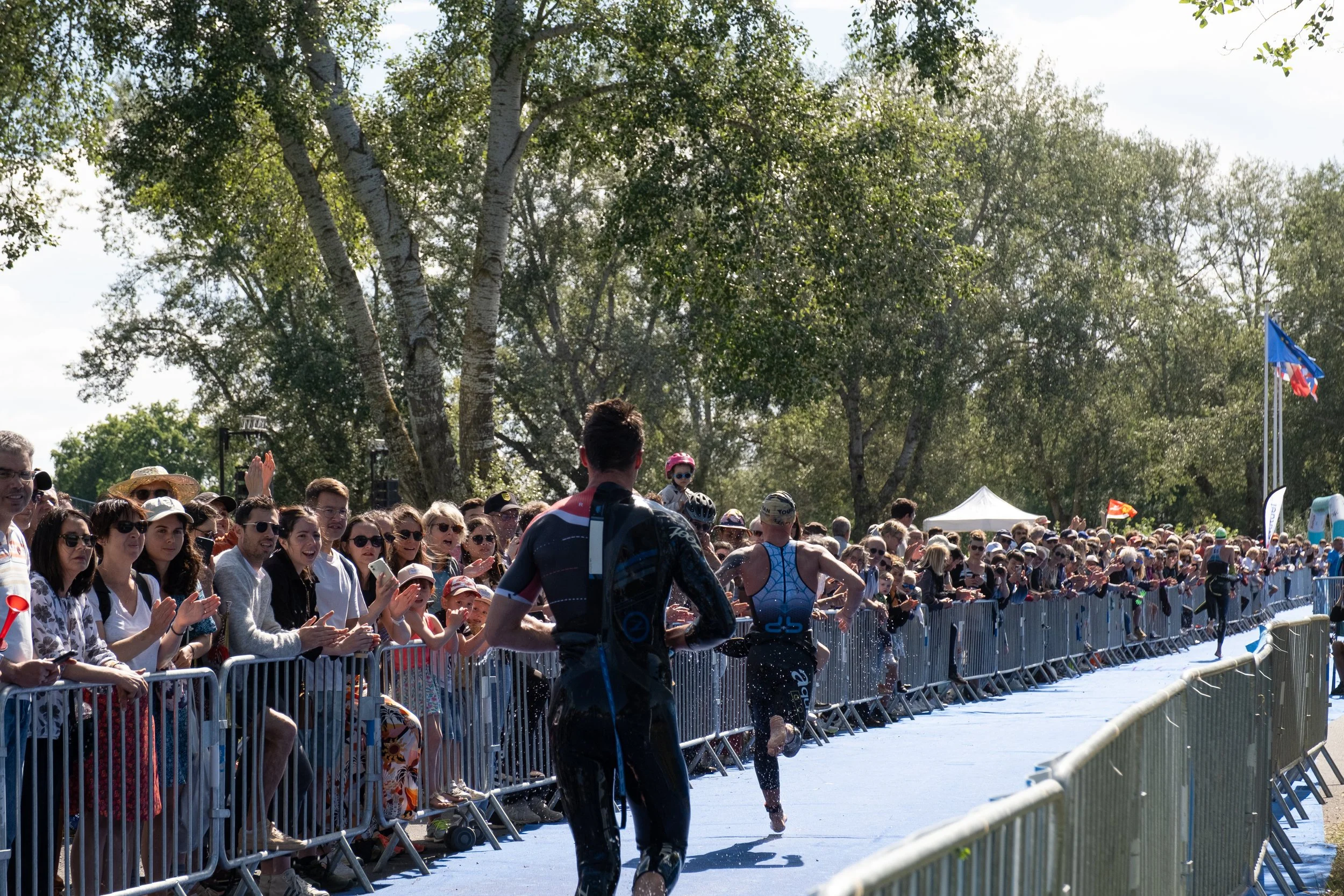 Des athlètes traversent la ligne d'arrivée d'un triathlon devant un public enthousiaste derrière une barrière de sécurité, avec des arbres en arrière-plan.