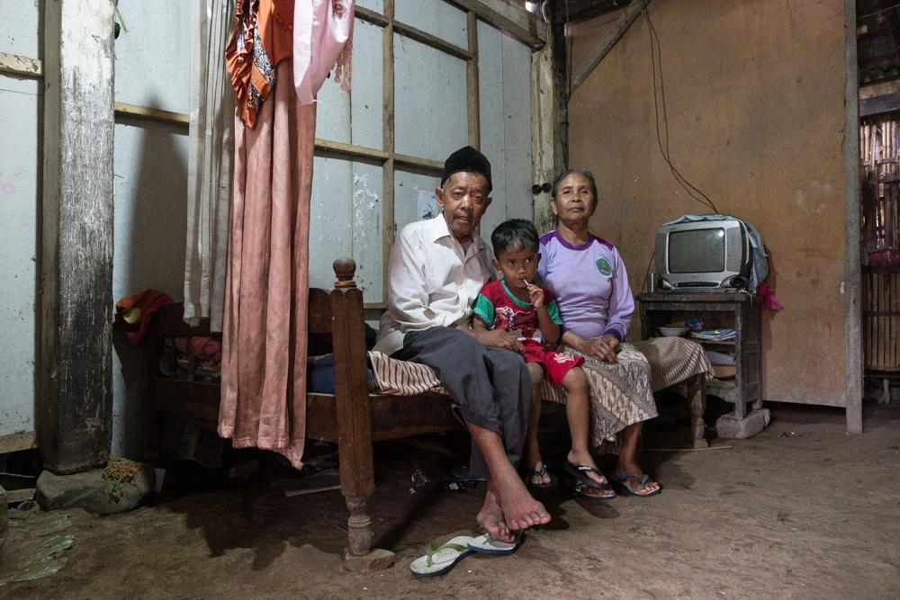 Une famille de quatre personnes assises dans une pièce simple, avec un vieux téléviseur et des vêtements suspendus en arrière-plan.