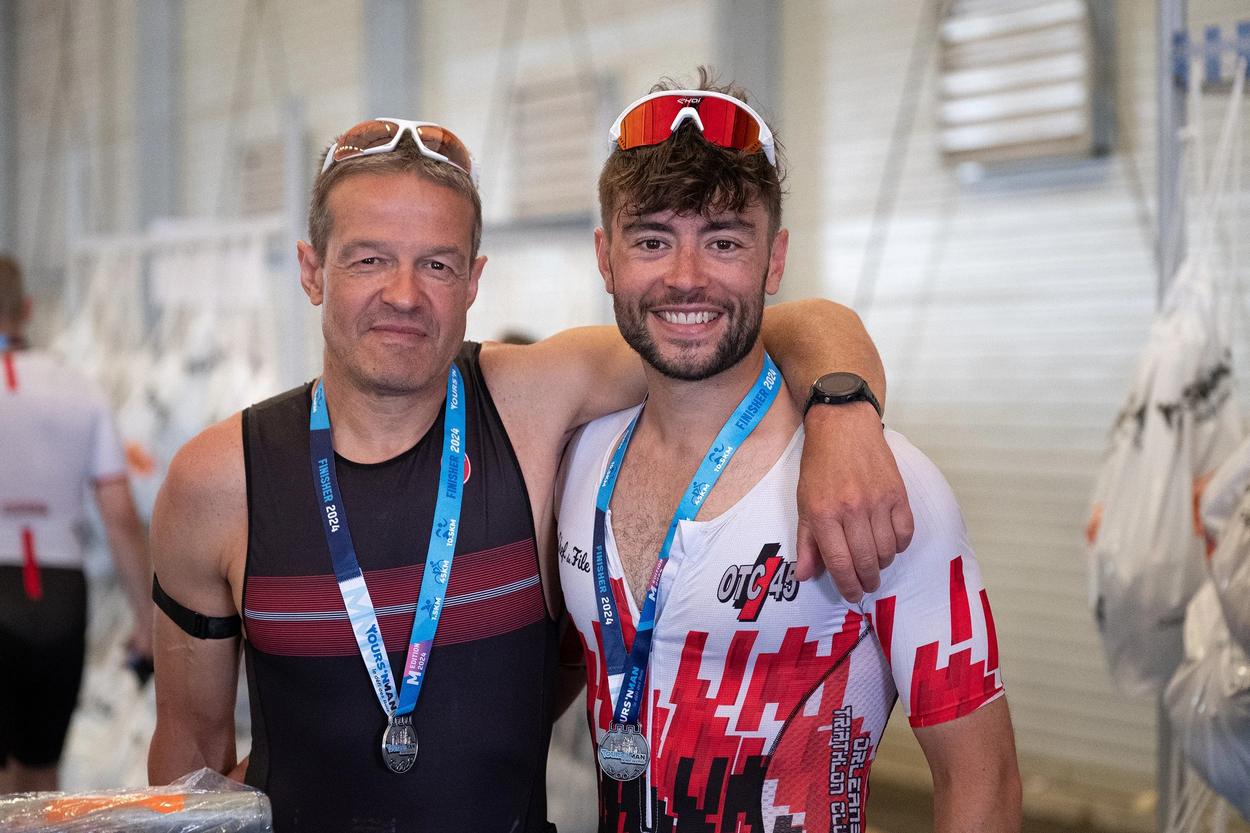 Deux hommes sportifs souriants, portant des médailles de finisher, se serrant l'épaule après une compétition, dans un environnement intérieur, avec d'autres personnes en arrière-plan.