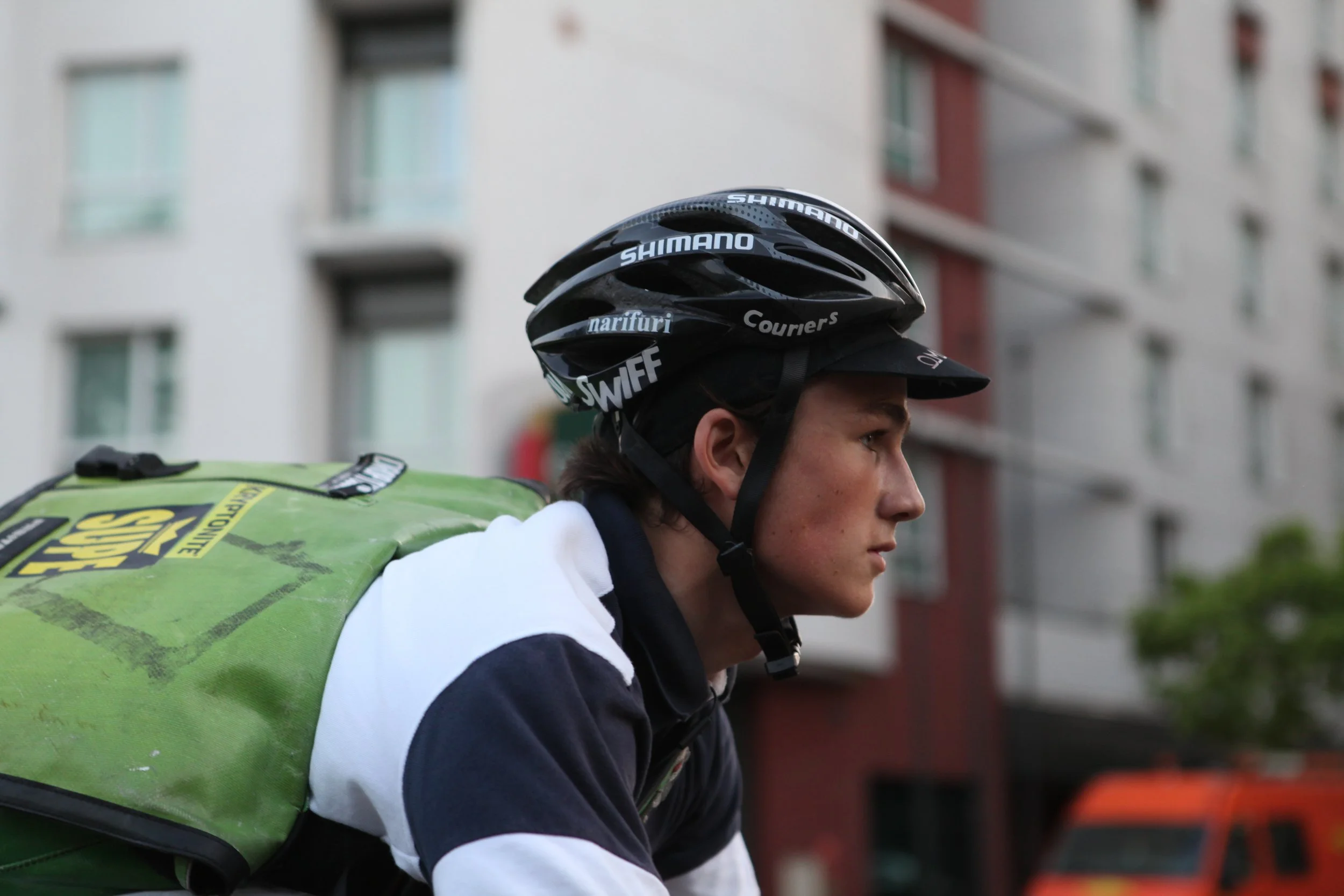 Jeune livreur à vélo portant un casque et un sac à dos vert dans une ville.