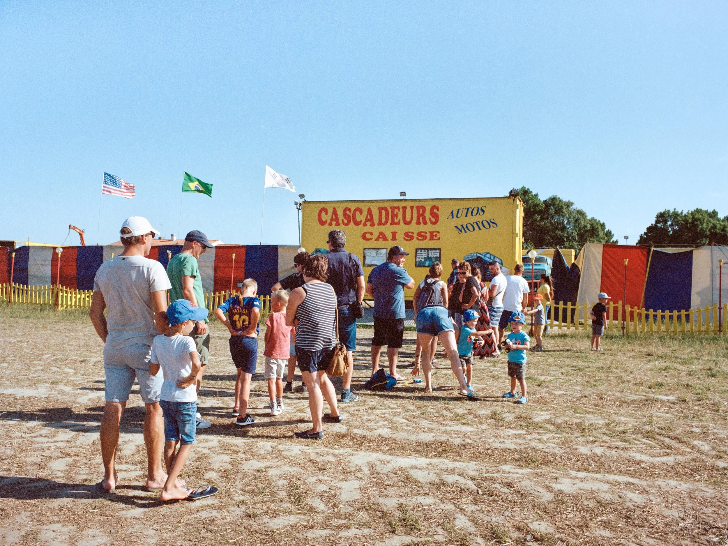 Une file d'attente d'enfants et d'adultes devant une billetterie de forains avec un panneau jaune marqué « Cascadeurs Caisse » et des drapeaux américains, brésiliens et sud-coréens en arrière-plan.