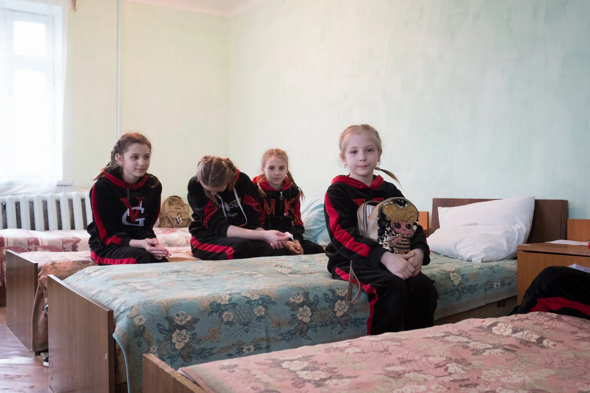 Quatre jeunes orphelines ukrainiennes assises sur des lits dans une chambre, portant des survêtements noirs et rouges, avec une des filles tenant un sac et un jouet L.O.L. dans ses bras.