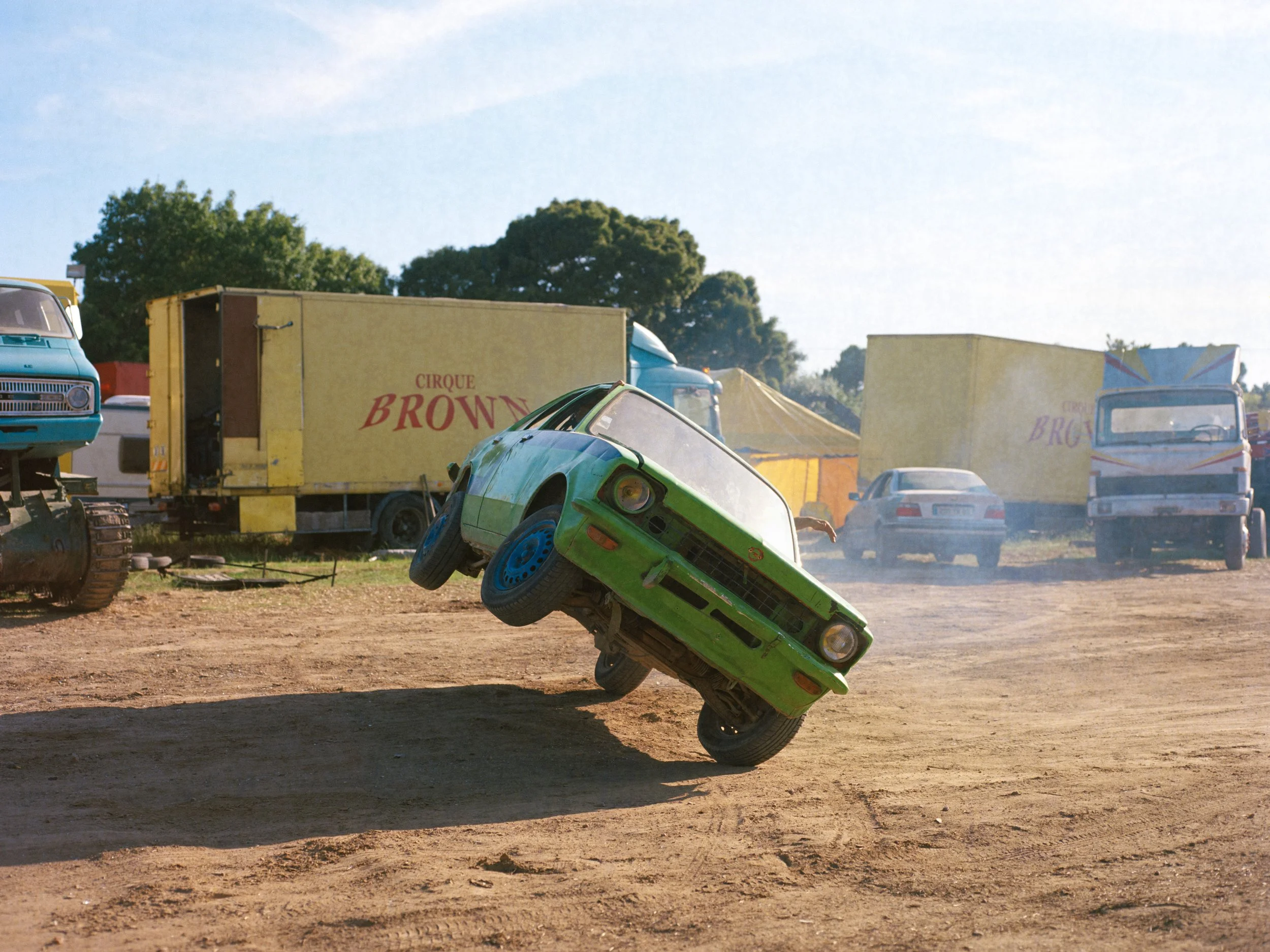 Une voiture verte effectuant une figure acrobatique lors d'un show de Monster Truck, avec des camions en arrière-plan portant l'inscription 'Cirque Brown'.