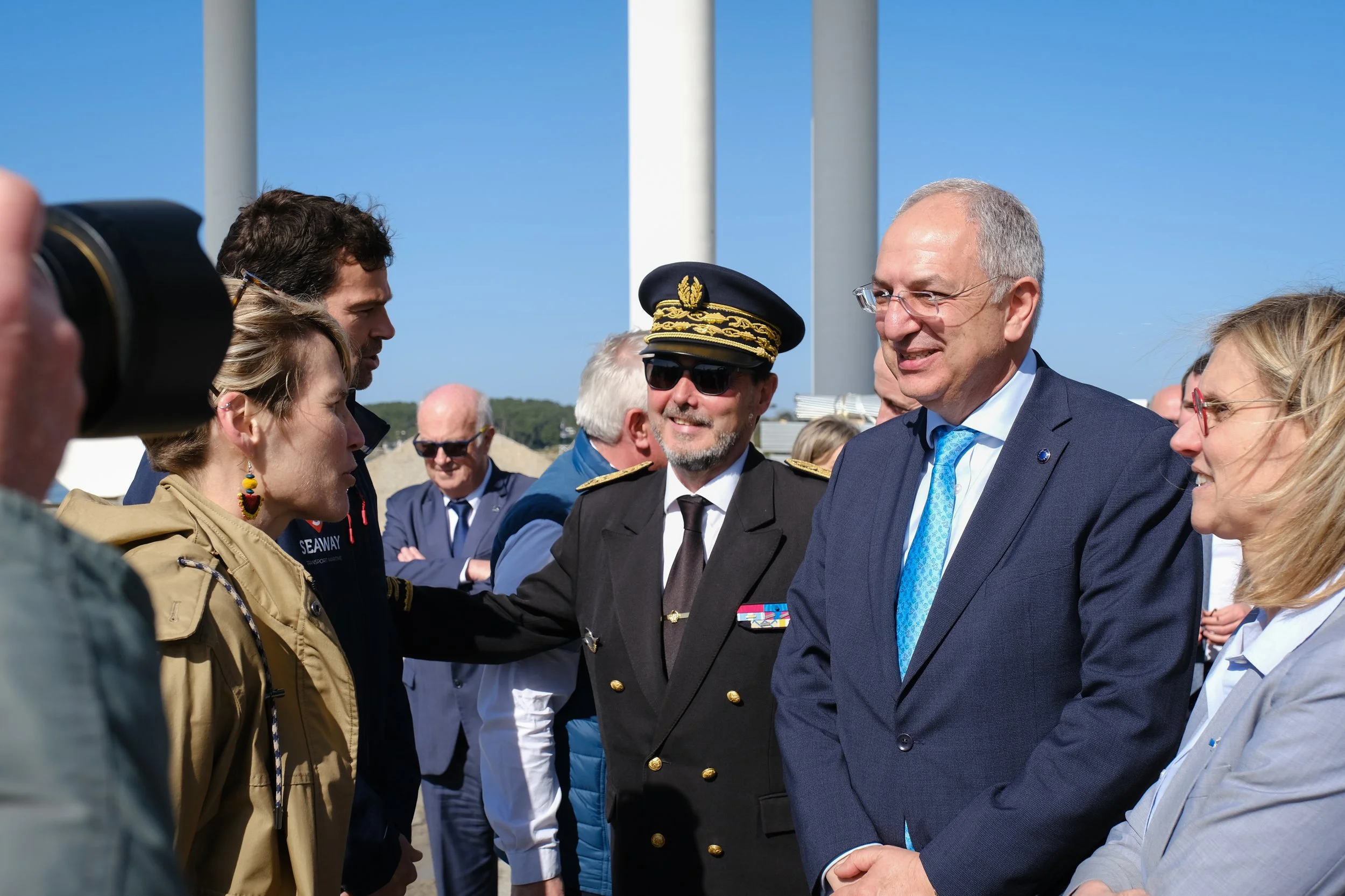 Groupe de personnes réunies en extérieur, dont un homme en uniforme militaire, un homme en costume avec une cravate bleue, une femme avec des lunettes, et d'autres personnes, en discussion lors d'un événement officiel.