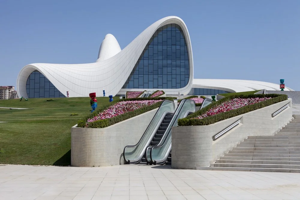 Centre Heydar Aliyev de Zaha Hadid à Baku, en Azerbaïdjan, une architecture moderne avec une forme incurvée, grandes fenêtres en verre, escaliers et escalators devant un jardin fleuri.