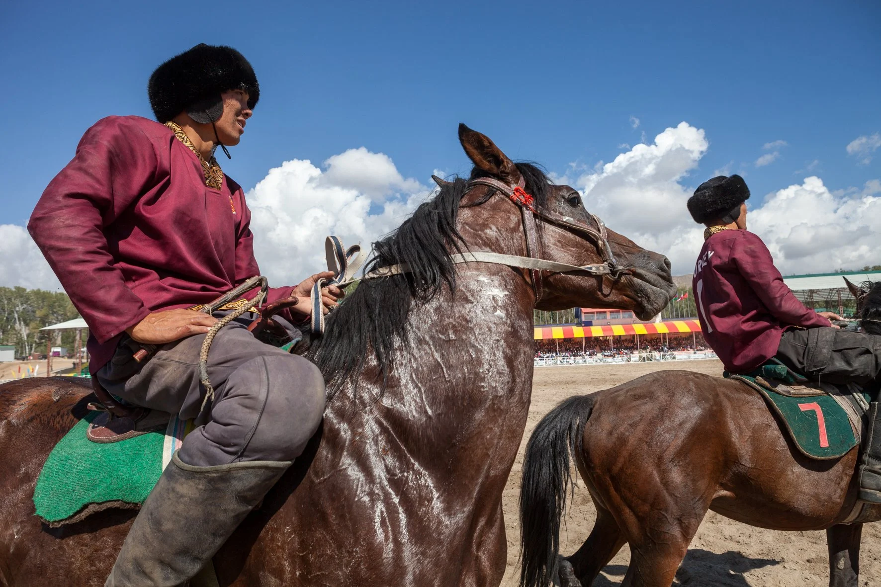 Deux jockeys à cheval lors d'une course, portant des vêtements bordeaux avec des chapeaux en fourrure. Le ciel est bleu avec des nuages blancs, et le public est visible en arrière-plan.