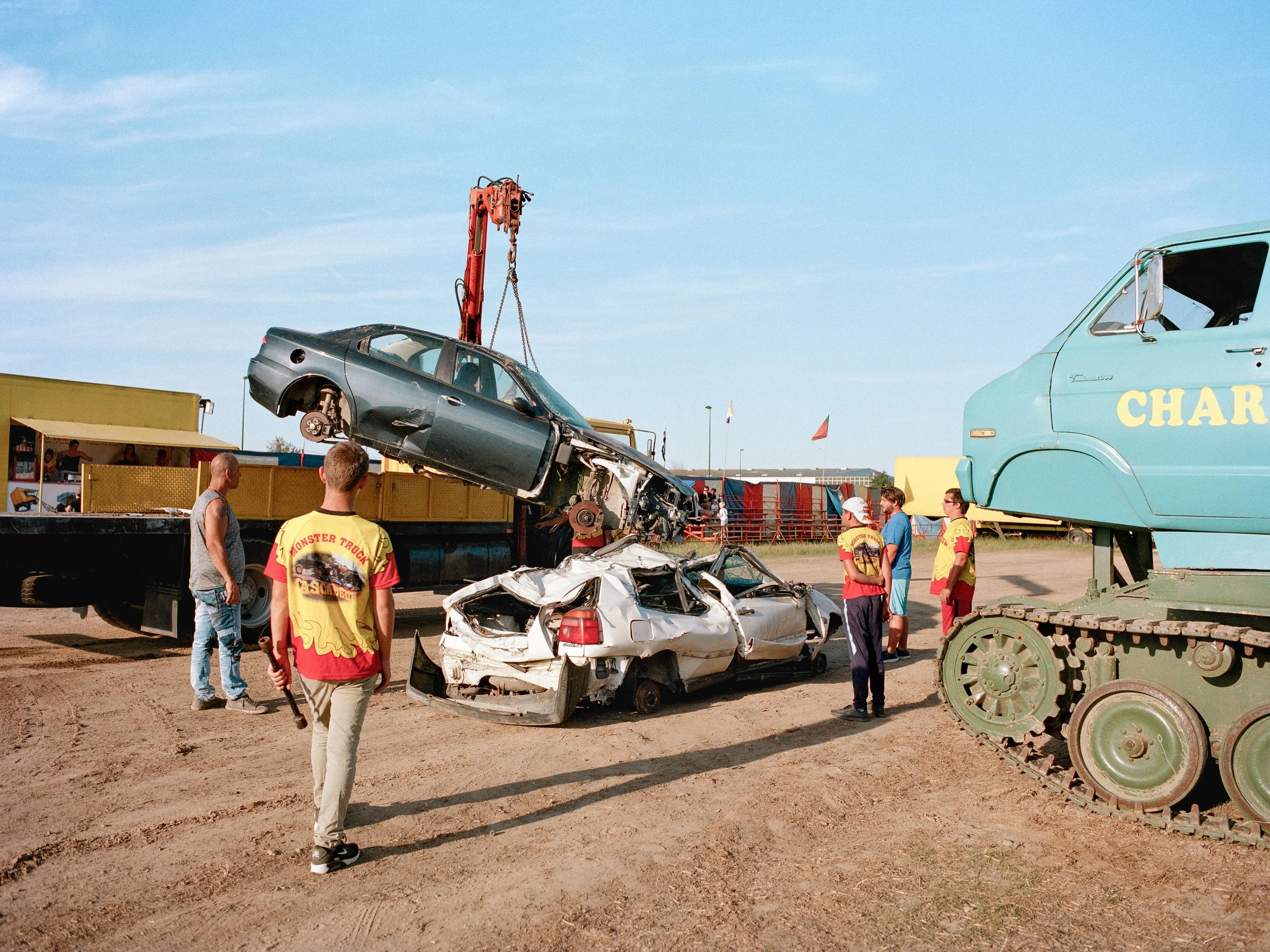 Fin d'un spectacle de destruction de voitures en plein air. Un engin de chantier remet les voitures accidentées sur un camion. Plusieurs personnes regardent.